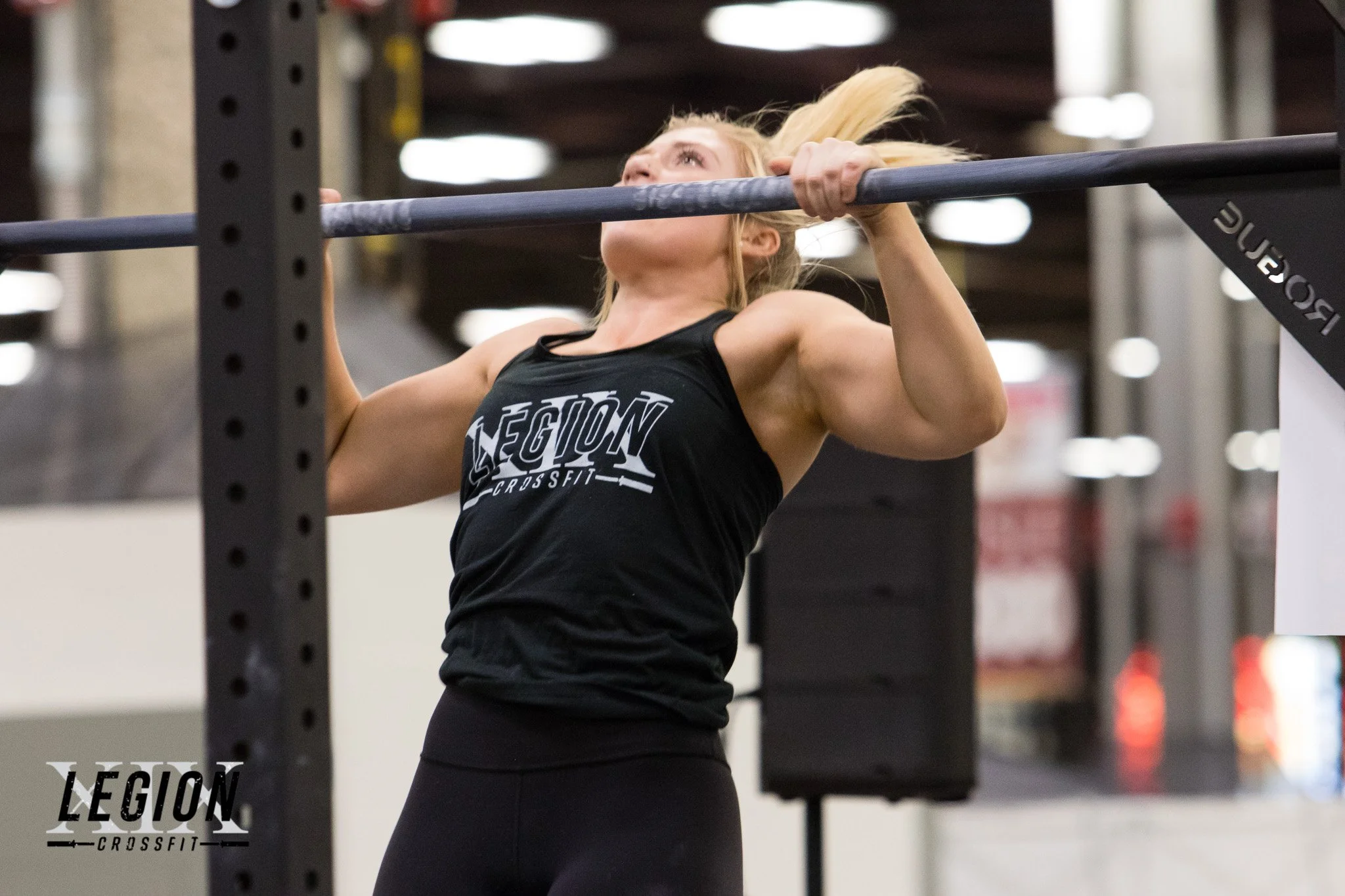 A woman with blonde hair lifting a barbell overhead at a gym, wearing a black tank top with 'LEGION CROSSFIT' printed on it.