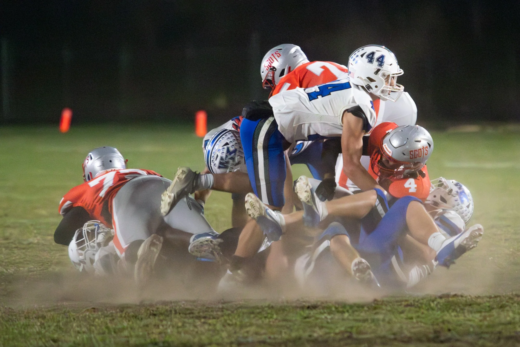 American football players in red, white, blue, and gray uniforms on a dirt field during a game at night, with multiple players tackling each other.