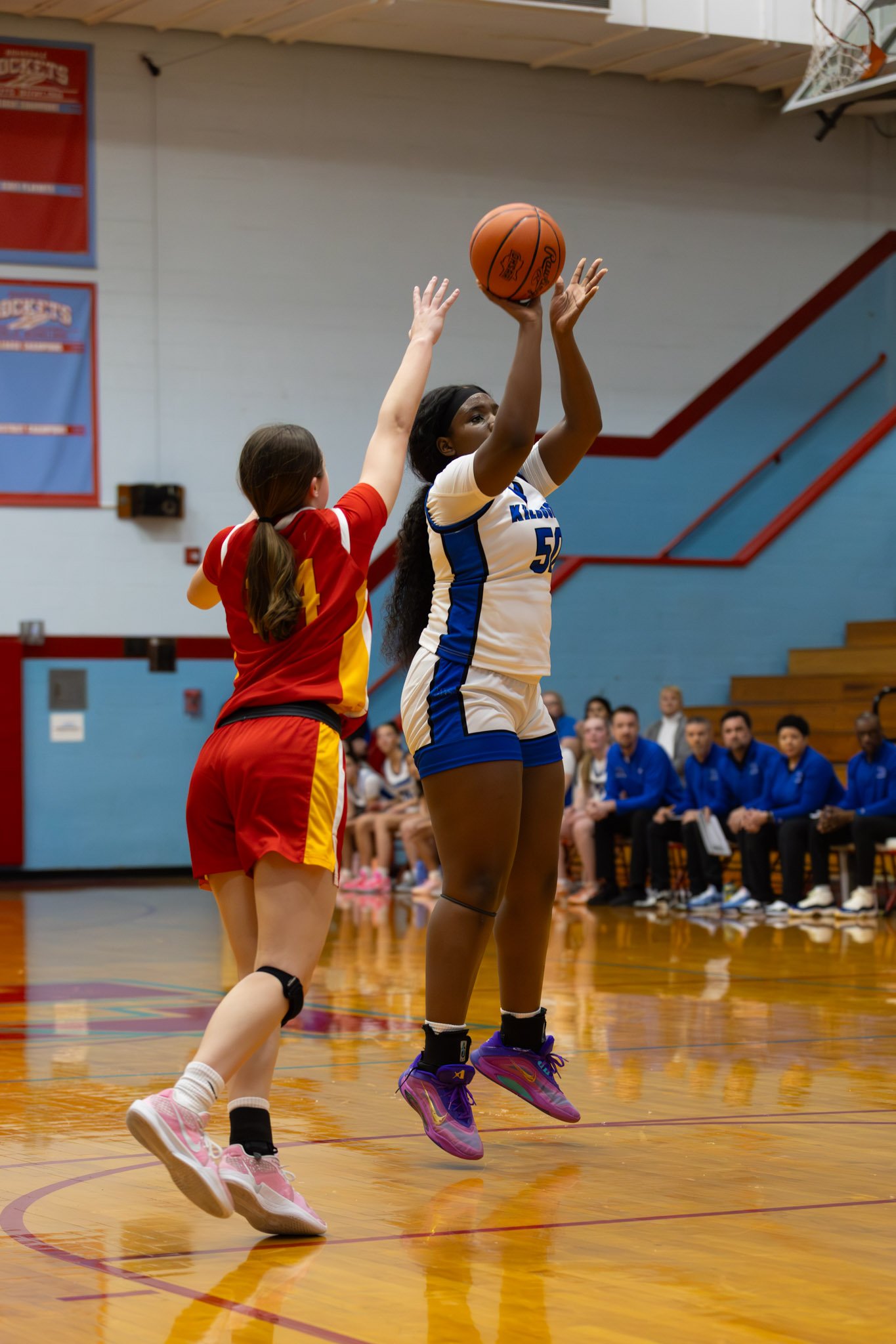 A female basketball player in a white and blue uniform jumping to shoot the ball while an opposing player in a red and yellow uniform attempts to block the shot during a game in a gymnasium.