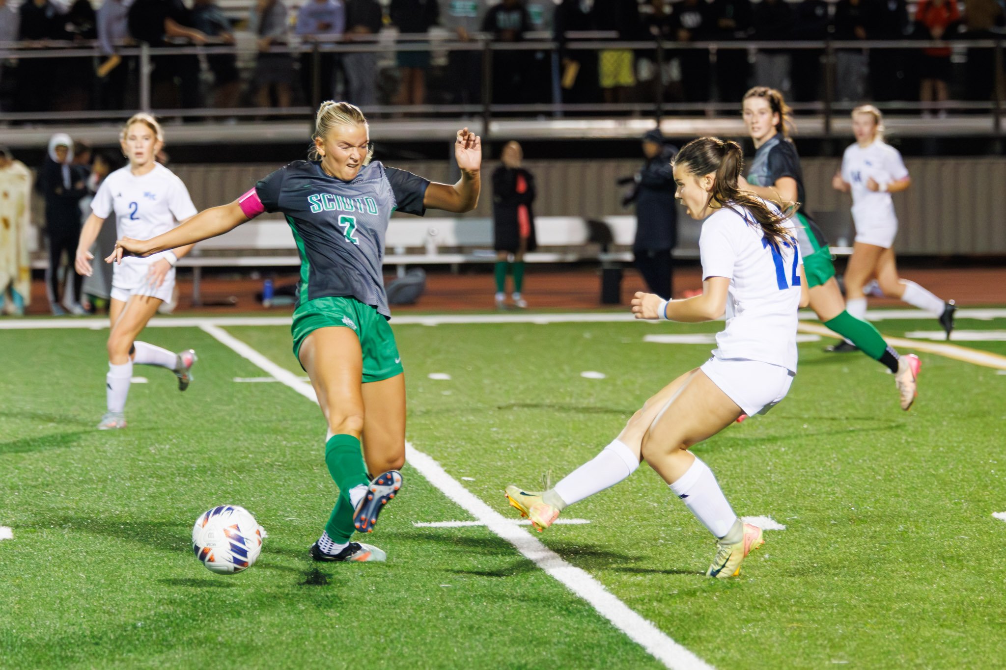 Two female soccer players from opposing teams compete for the ball on the field during a match, with other players and spectators in the background.