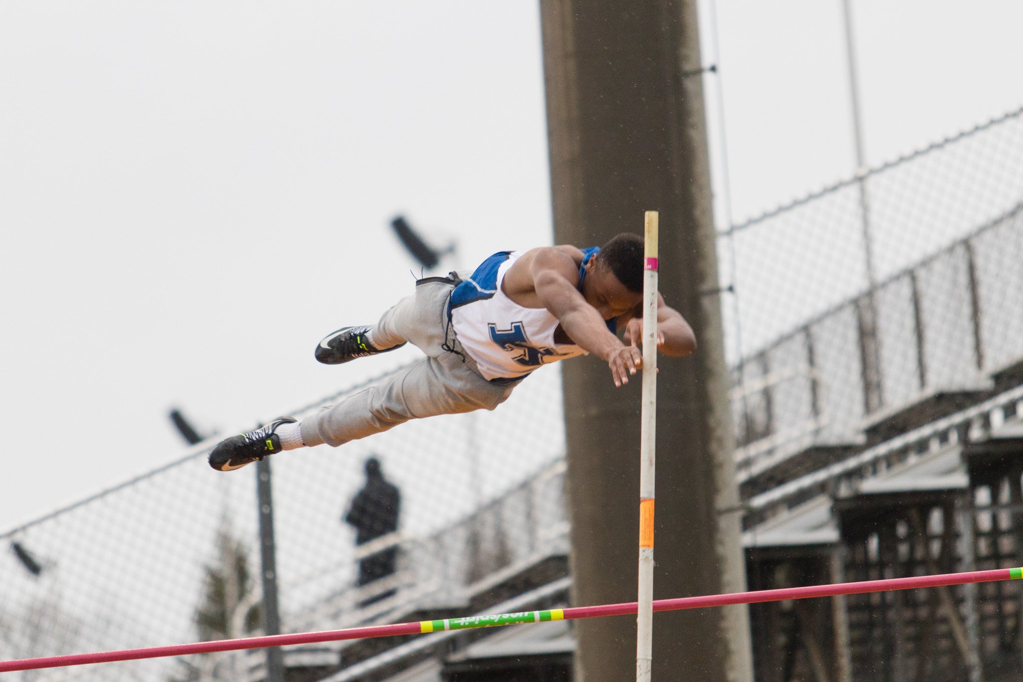 An athlete in a white and blue uniform is mid-air during a pole vault attempt, clearing a horizontal bar on an outdoor track field.