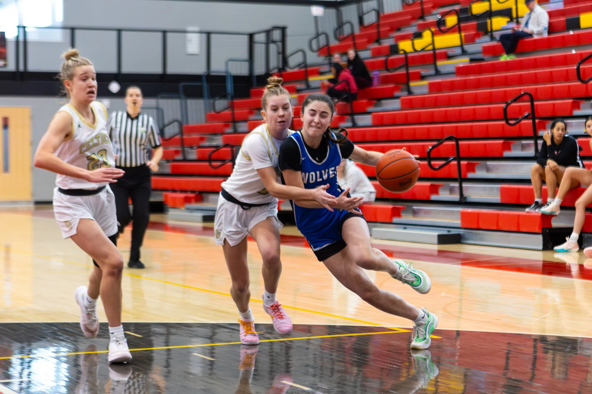 A women's basketball game in a gymnasium showing two players fighting for the ball while another player and a referee look on. Red bleachers with spectators are in the background.