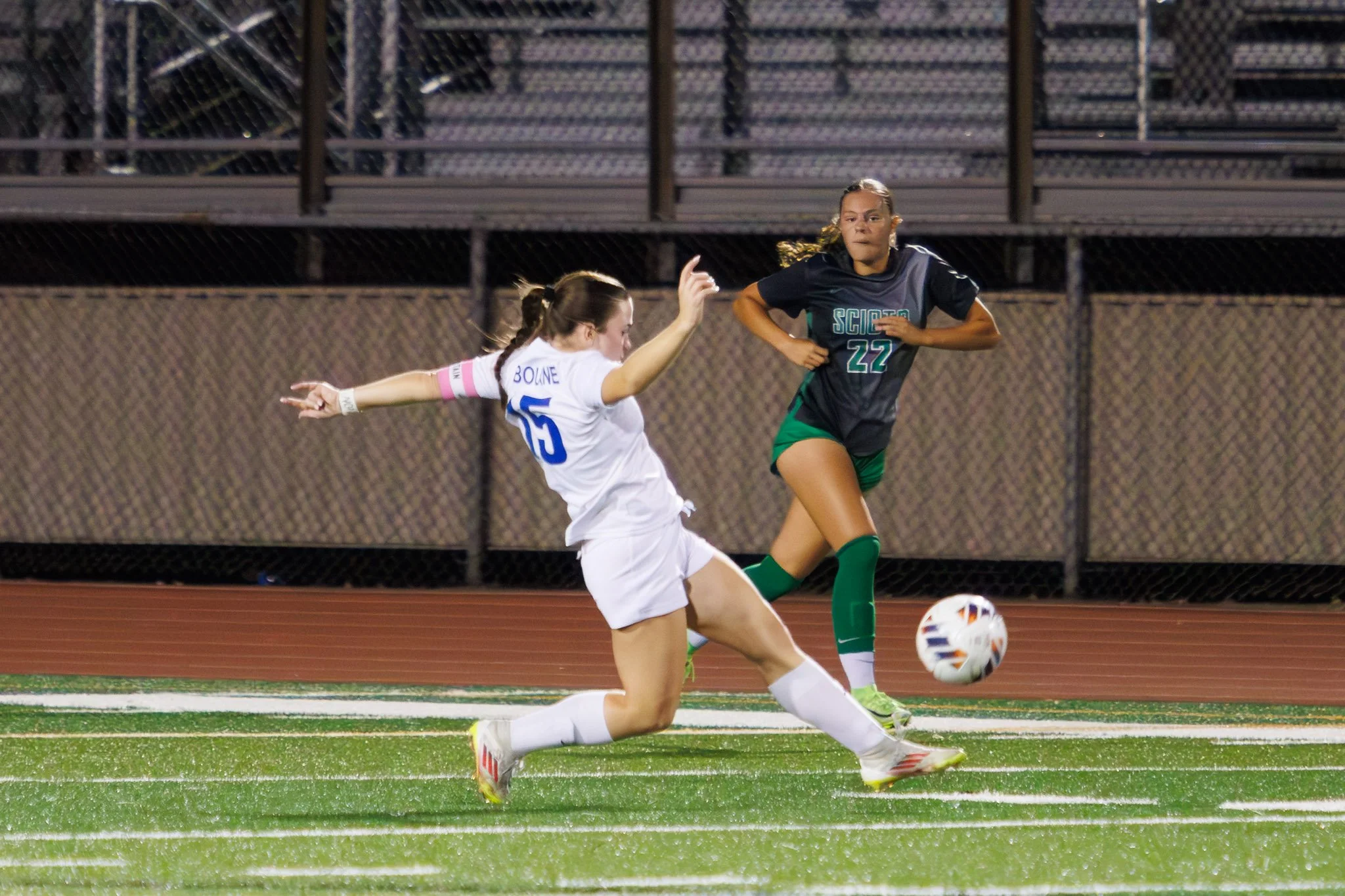 Two female soccer players competing for the ball on a field at night, with one in a white uniform and the other in a black and green uniform.