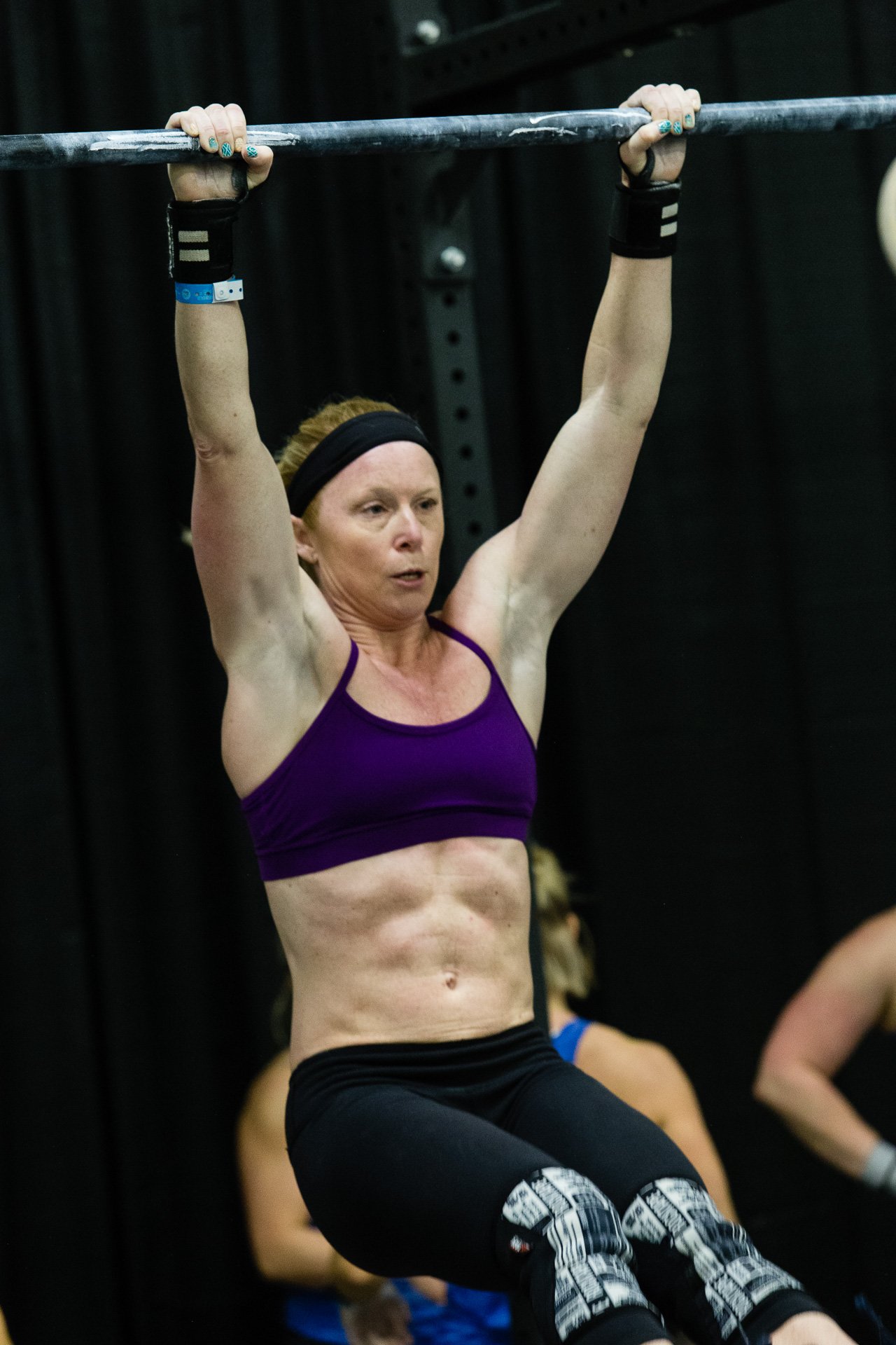 A woman with blonde hair, wearing a black headband, purple sports bra, and black leggings, is lifting herself up with her arms in a fitness or CrossFit gym. 