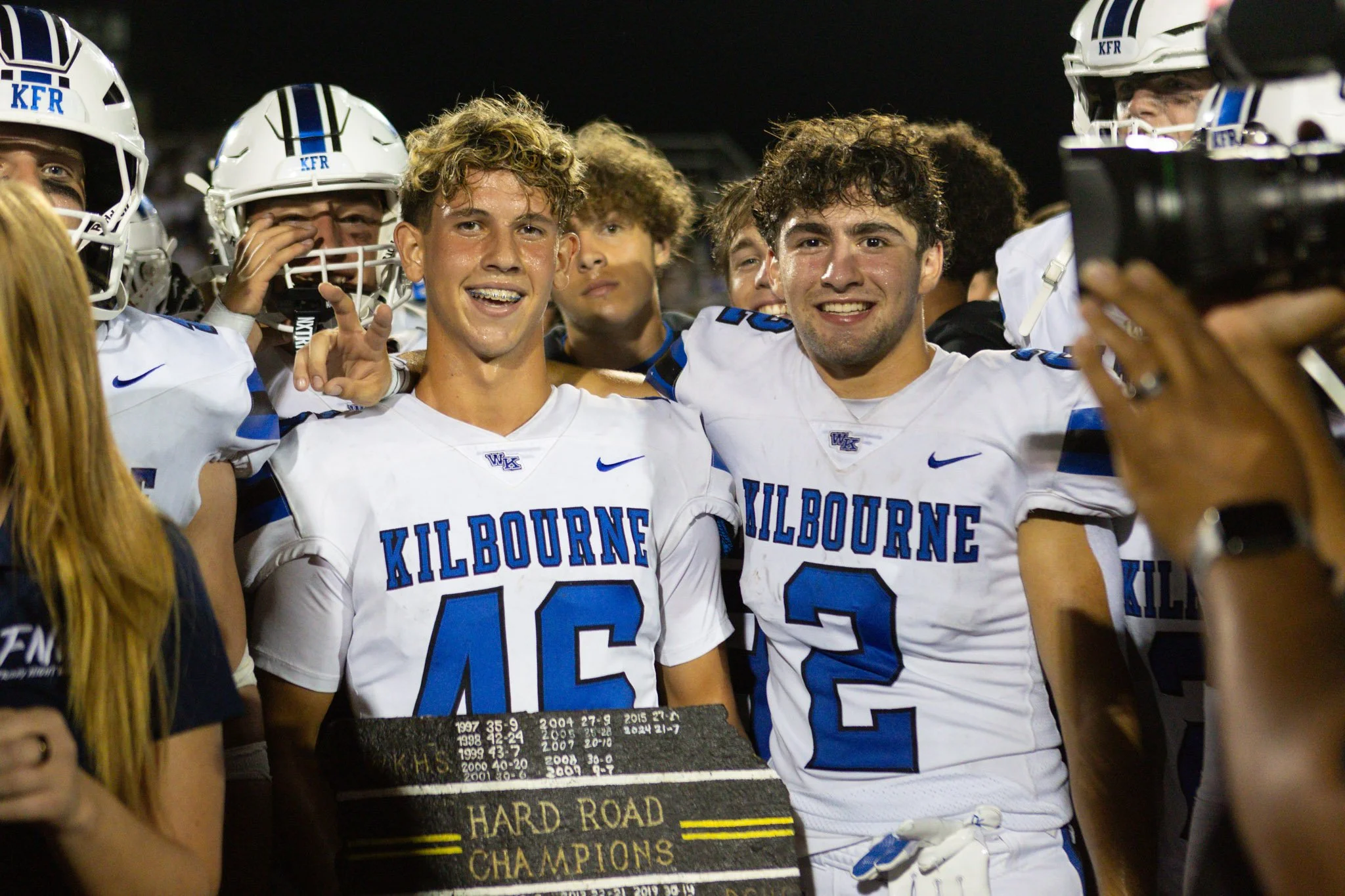Two football players from Kilbourne High School celebrating their victory, holding a trophy that says "Hard Road Champions," surrounded by teammates and photographers at night.