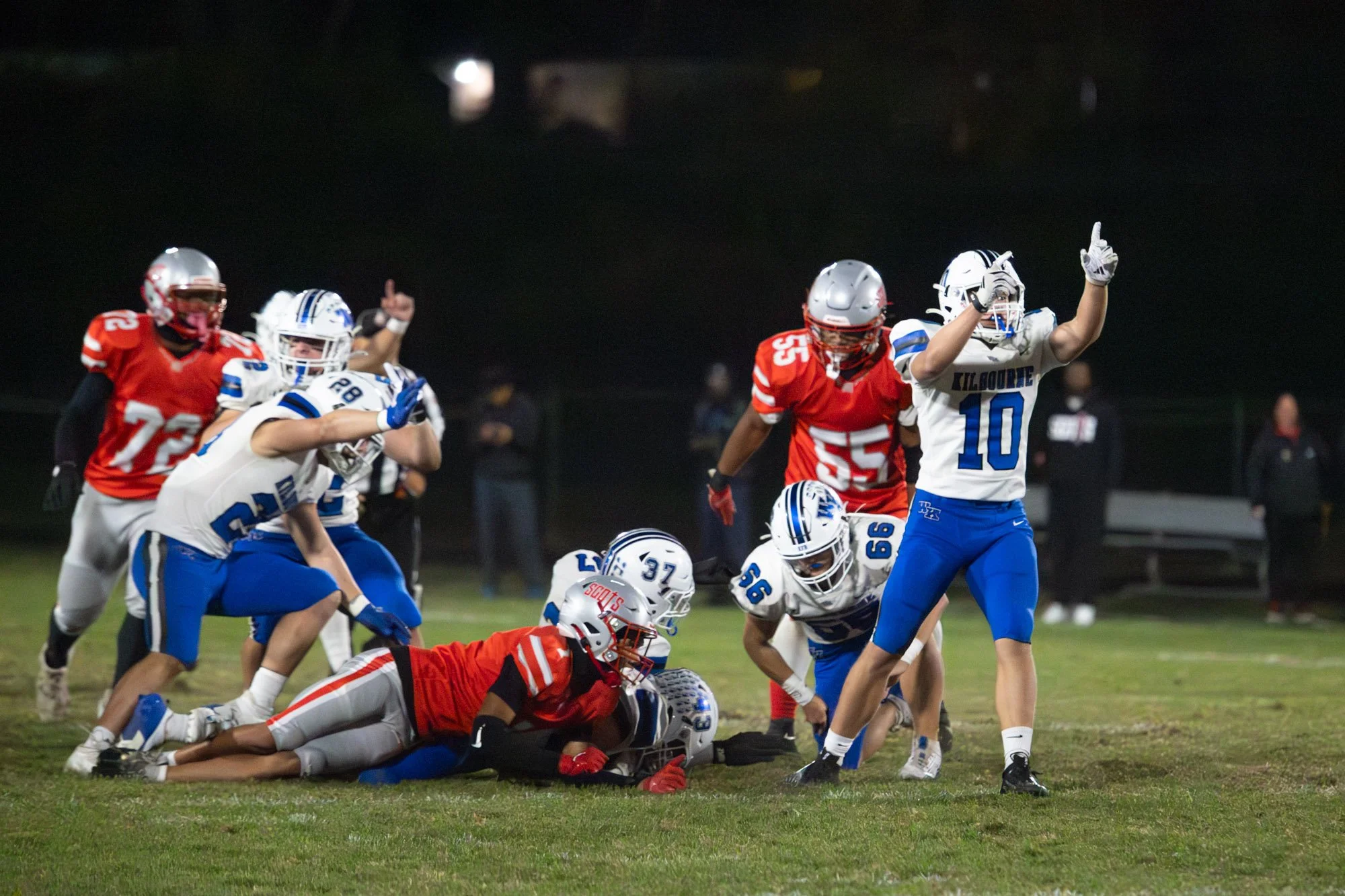 A nighttime football game with players in red and white and blue uniforms. Players are on the field, some on the ground, others standing, with one player in a white uniform raising her hand.