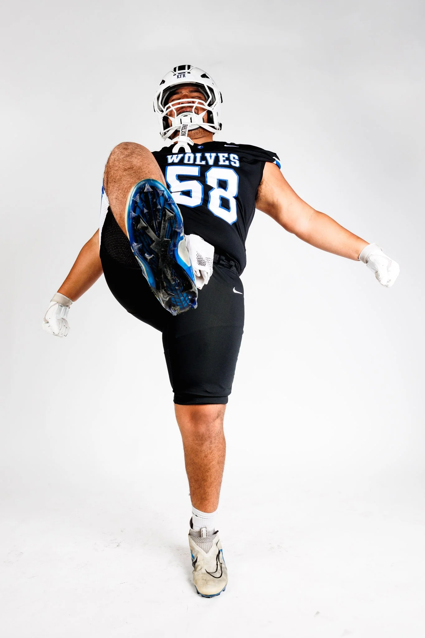 From a media day shoot, a football player in a black jersey with the number 58 and 'WOLVES' written on it, lifting one leg in a kicking pose, wearing a helmet, gloves, and cleats against a plain white background.