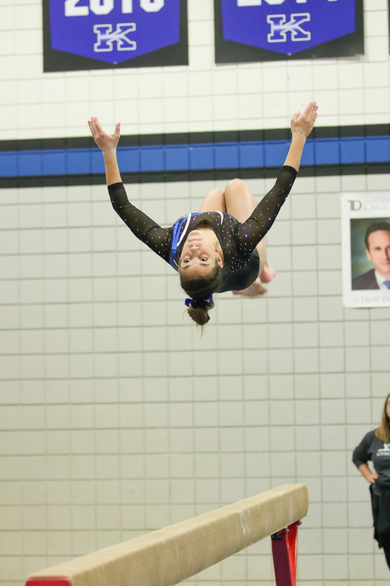 A young female gymnast dressed in a black leotard with sparkles is mid-air during a gymnastics routine, upside down, in a gymnasium with banners and a person standing in the background.