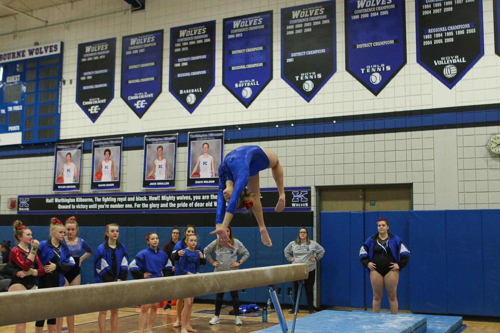 A female gymnast in a blue leotard performs a vault maneuver onto the vaulting table in a gymnasium, with spectators and fellow gymnasts watching. The gym has banners and posters on the walls.