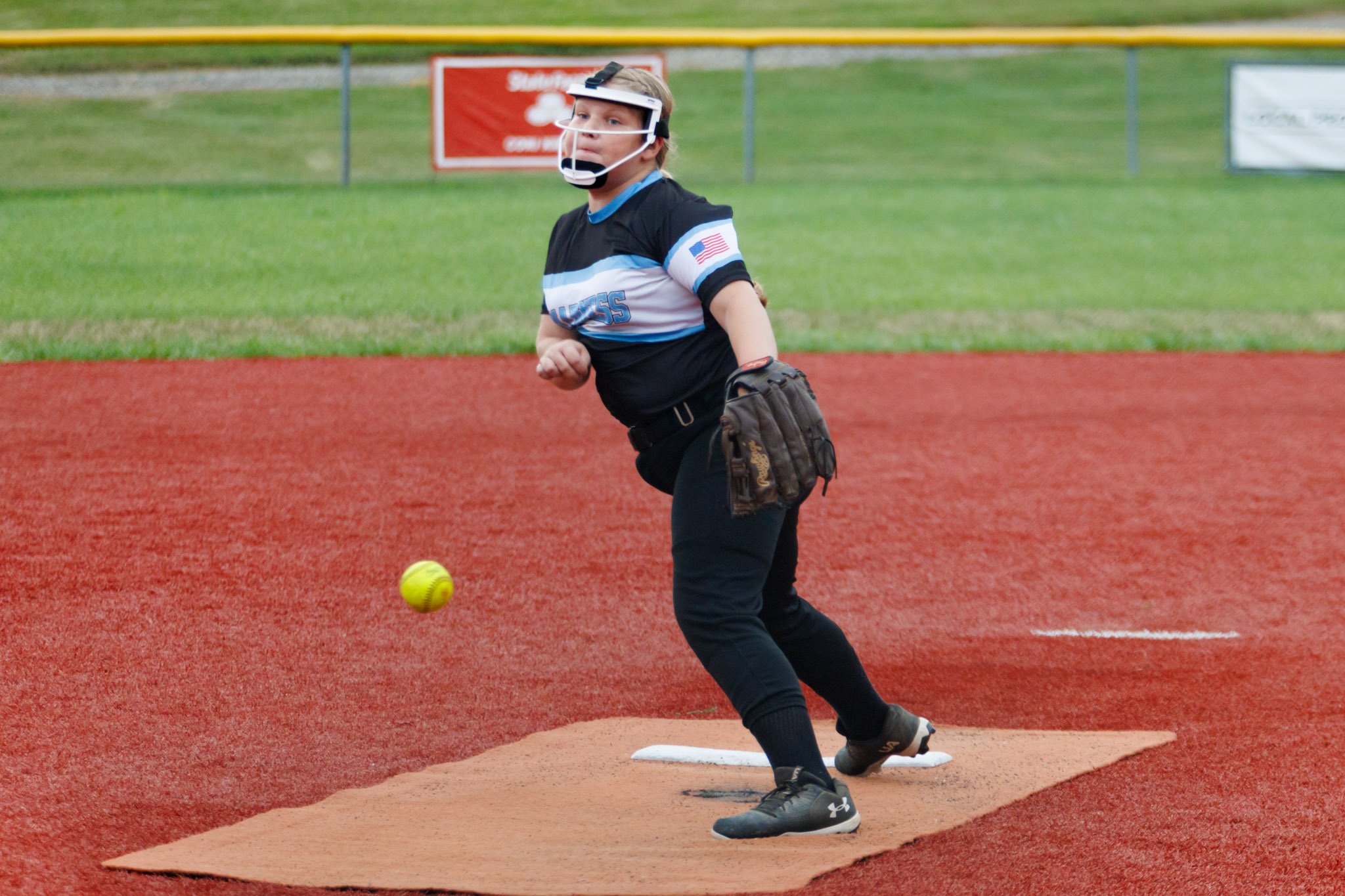 A young girl pitching softball on a field, wearing a face shield, black uniform, and holding a glove.
