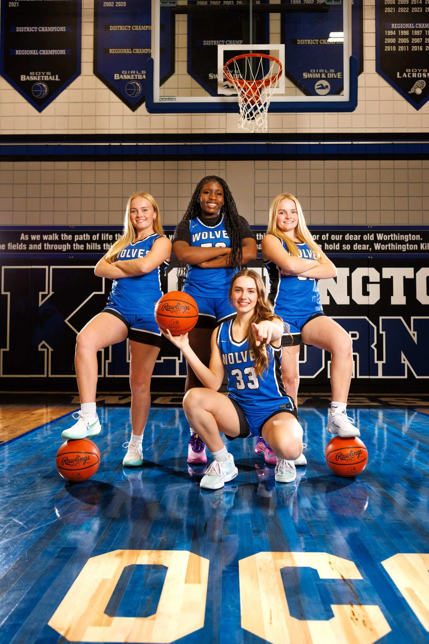 Media Day shoot Four female basketball players in blue jerseys posing in a gymnasium with a basketball court. One kneeling in front pointing at the camera, two standing with arms crossed behind, and one standing with arms crossed holding a basketball