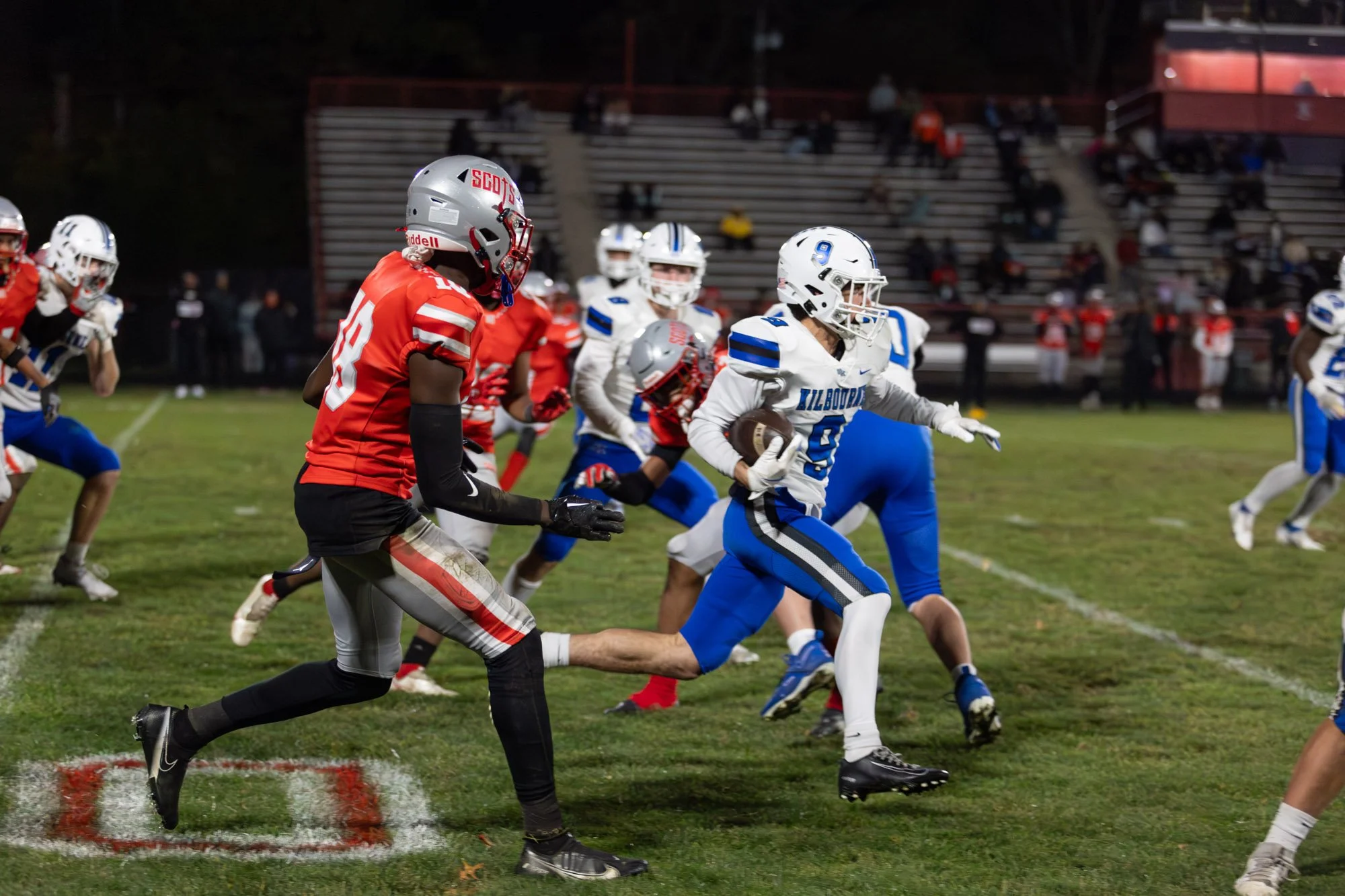 A football game in progress, with players from two teams running on the field. One team is wearing white and blue uniforms, while the other team is in red and black. The player in white and blue is holding a football and running, with other players c