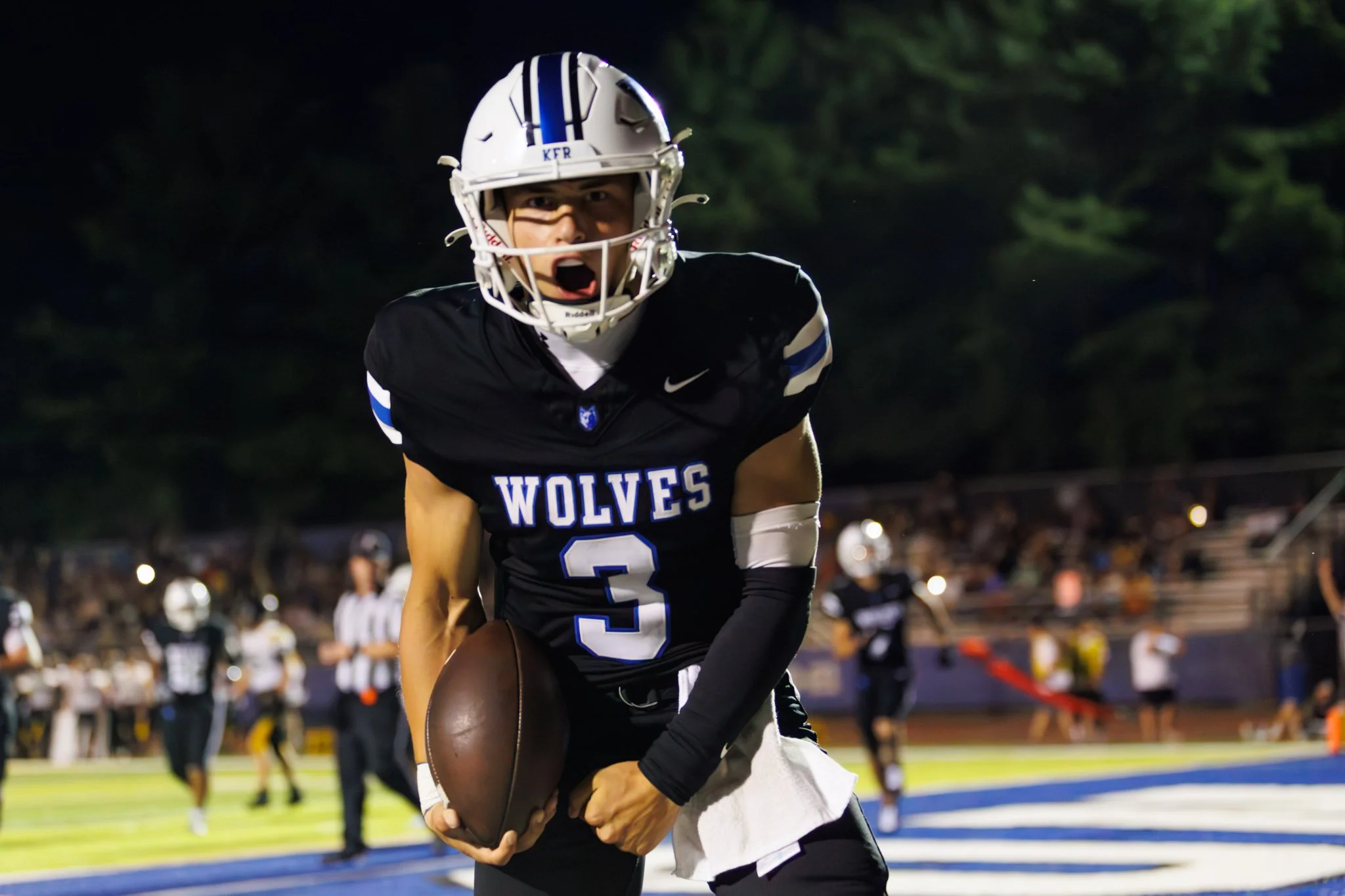 A football player in a black jersey with blue and white accents celebrating on the field, holding a football, wearing a white helmet with a blue stripe, during a game at night with spectators in the background.