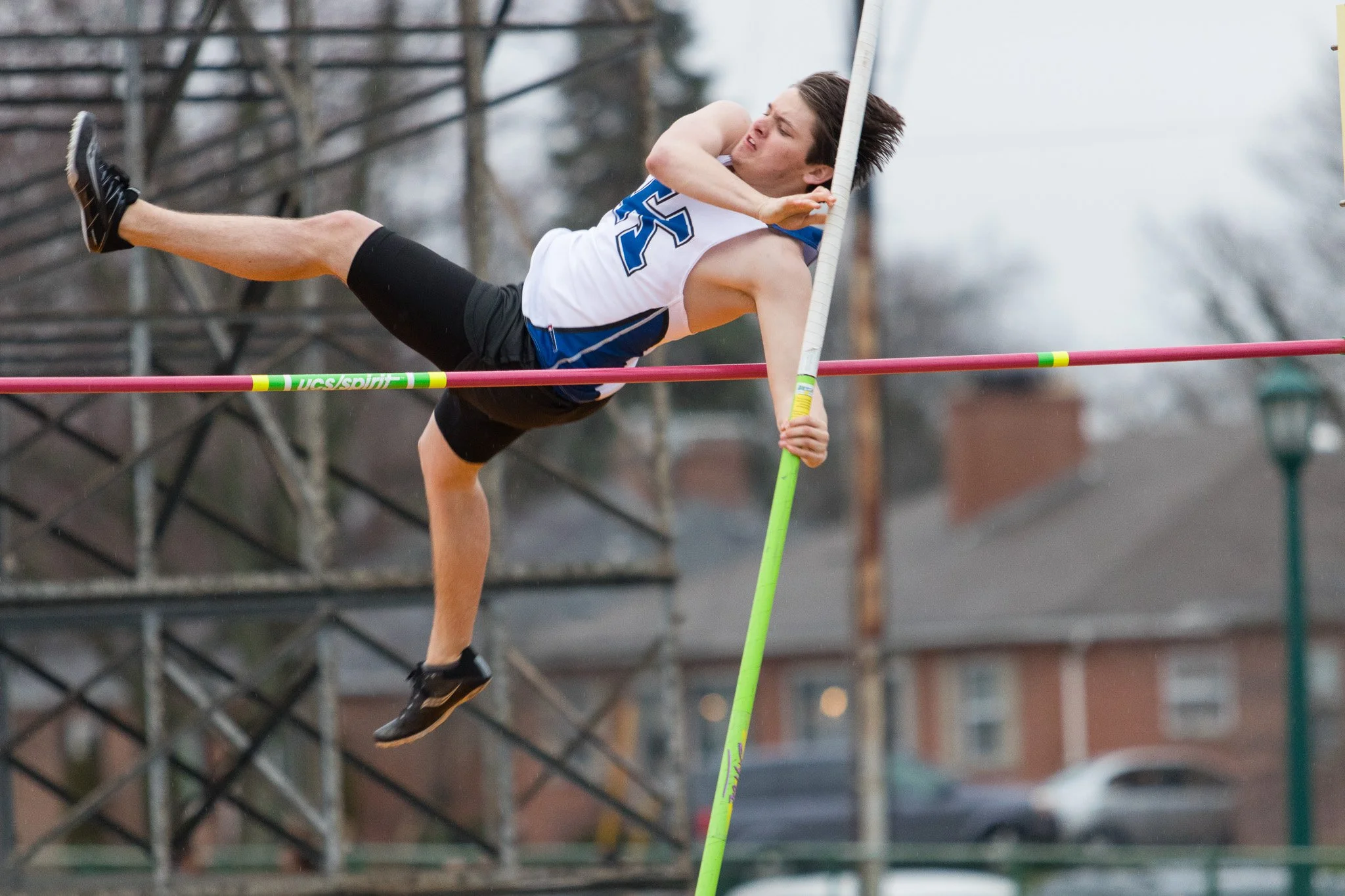A male pole vaulter clearing the high jump bar during a competition outdoors on a cloudy day.