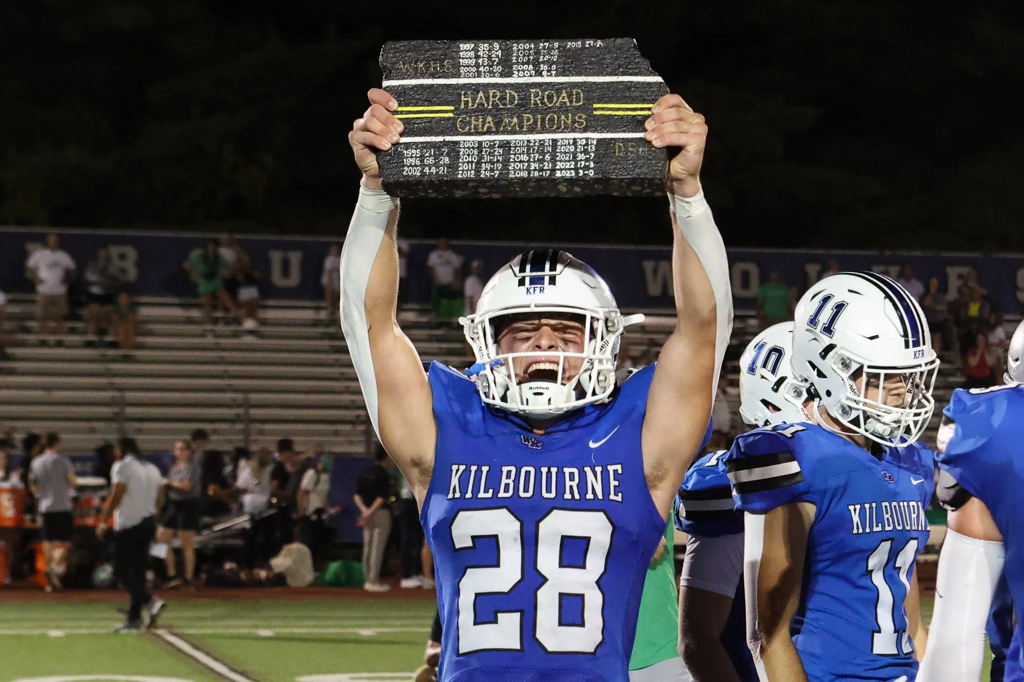 A football player in blue uniform with the number 28 and 'KILBOURNE' on the jersey holding a wooden sign with the words 'HARD ROAD CHAMPIONS' above his head. He is celebrating on a football field at night, with other players and spectators in the bac
