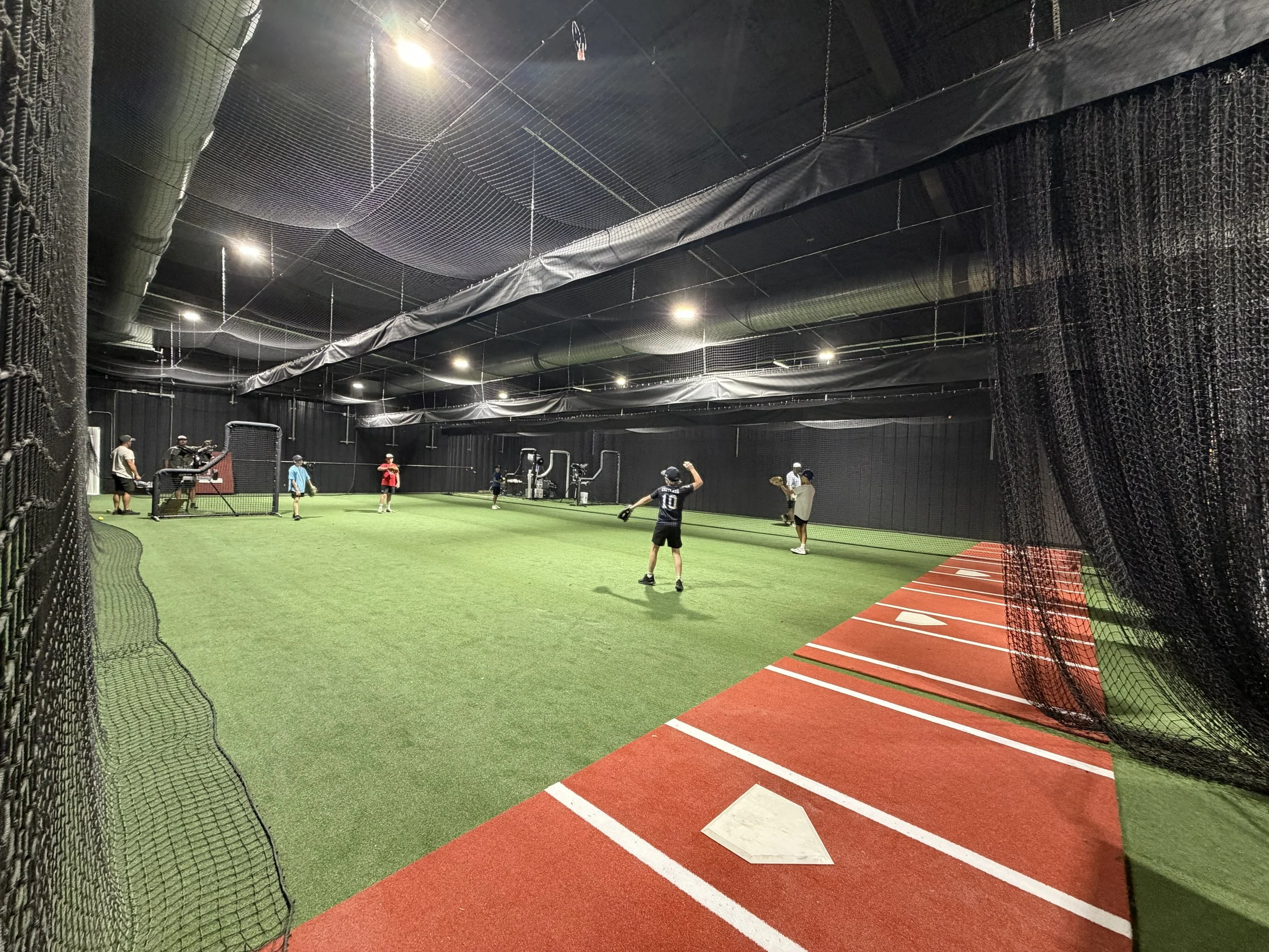Indoor baseball or softball training facility with netted fences, artificial turf, and several children and adults practicing batting and pitching.