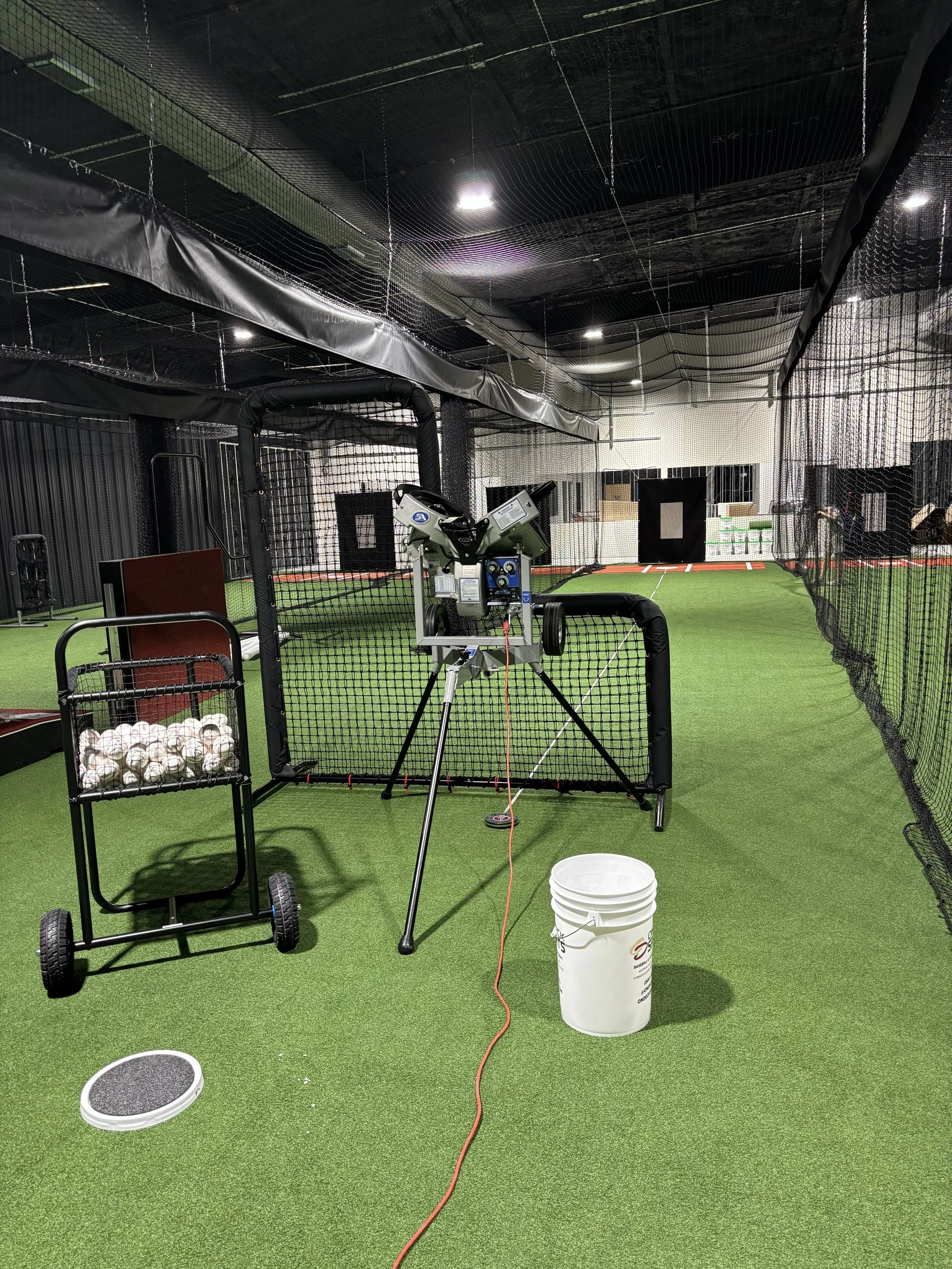 Indoor baseball practice area with netting, artificial turf, and baseballs in a cart, a bucket, and a pitching machine.