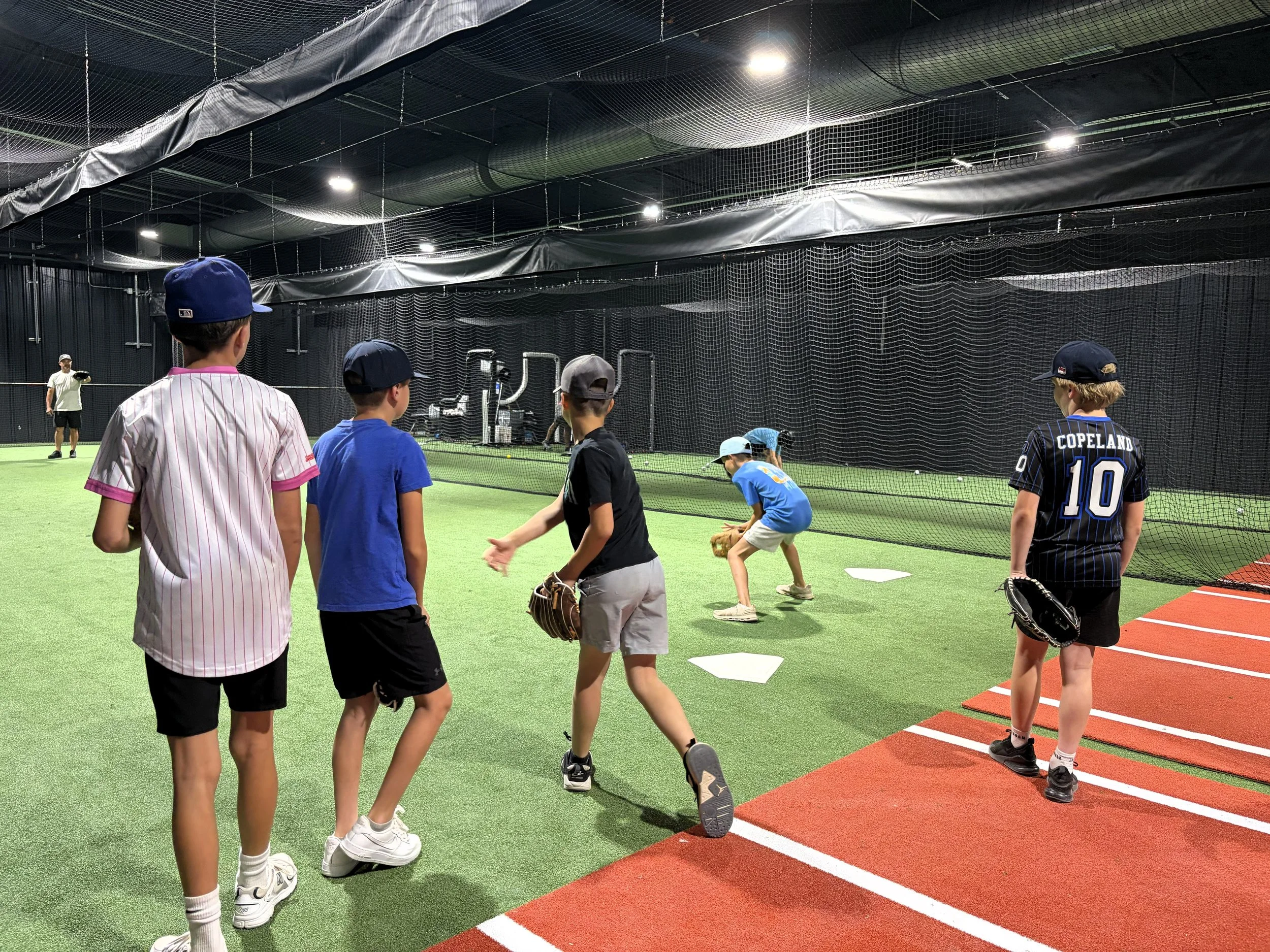 A group of young boys practicing baseball inside a batting cage with black netting, artificial turf, and white bases. They are wearing baseball caps and gloves, and are standing near the red running track.