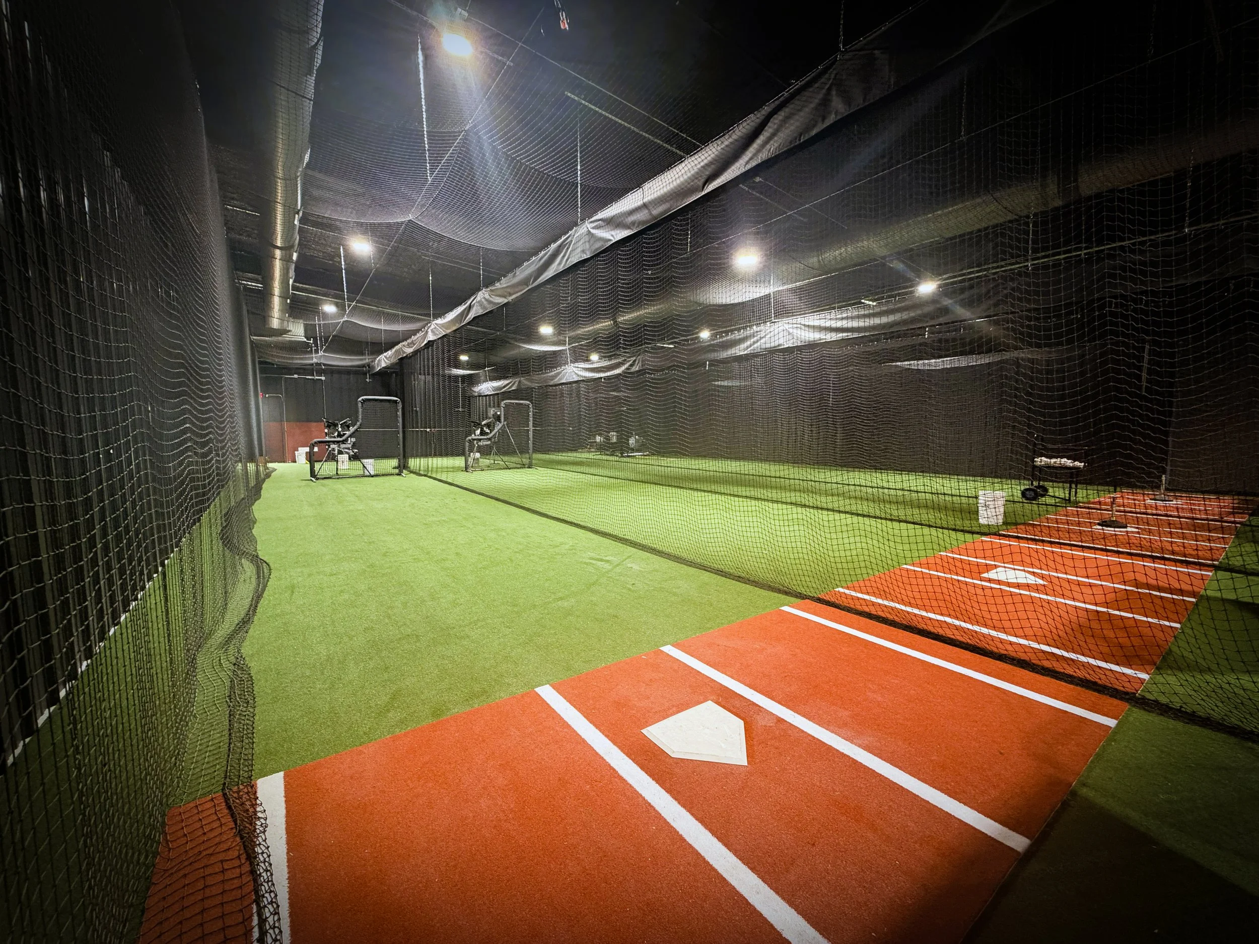 Indoor batting cage with artificial turf, netting surround, and baseball or softball target markings.