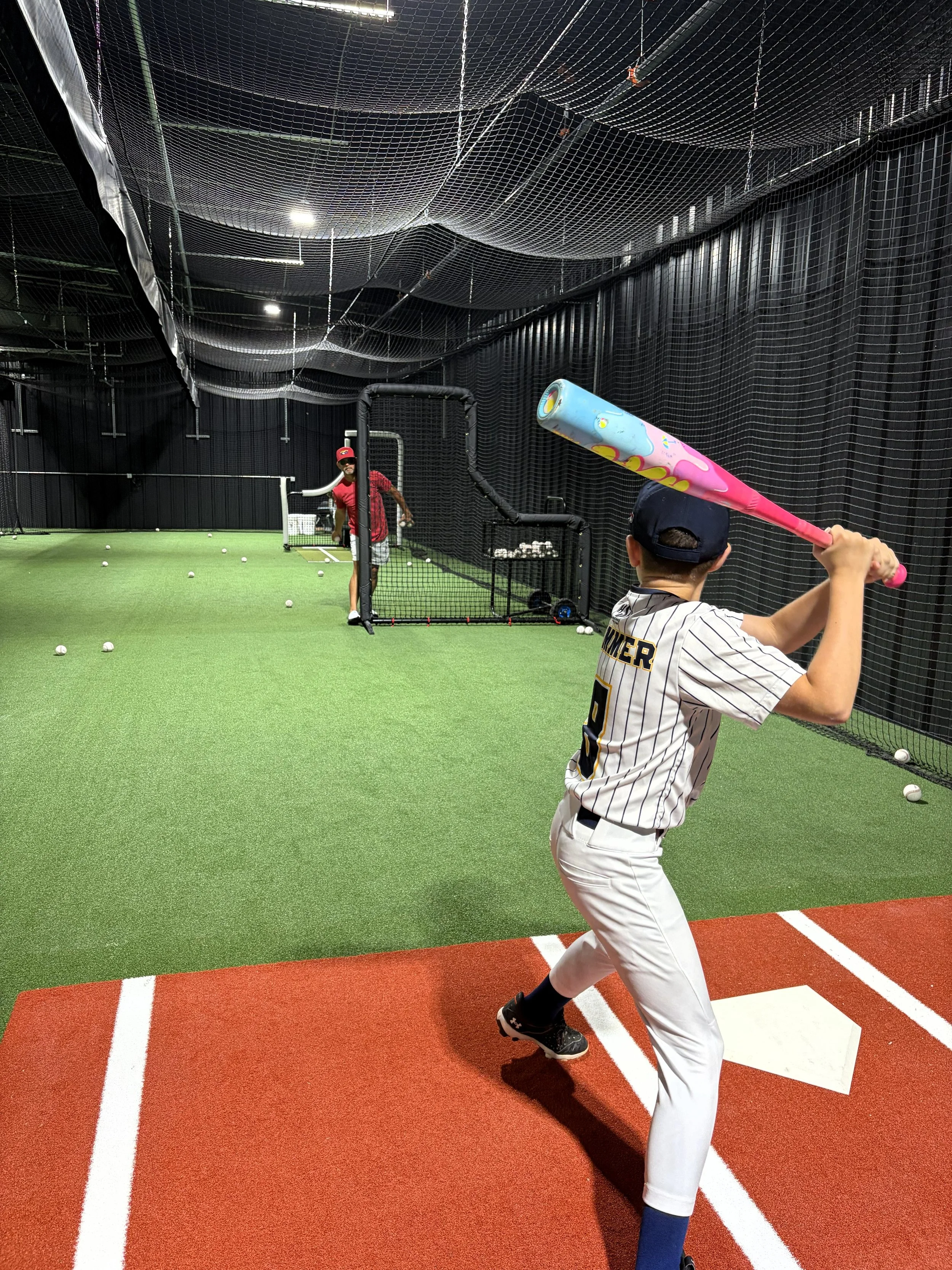 A young boy in baseball uniform practicing batting with a bat in a batting cage. An adult coach or trainer is standing in the background near a pitching machine.