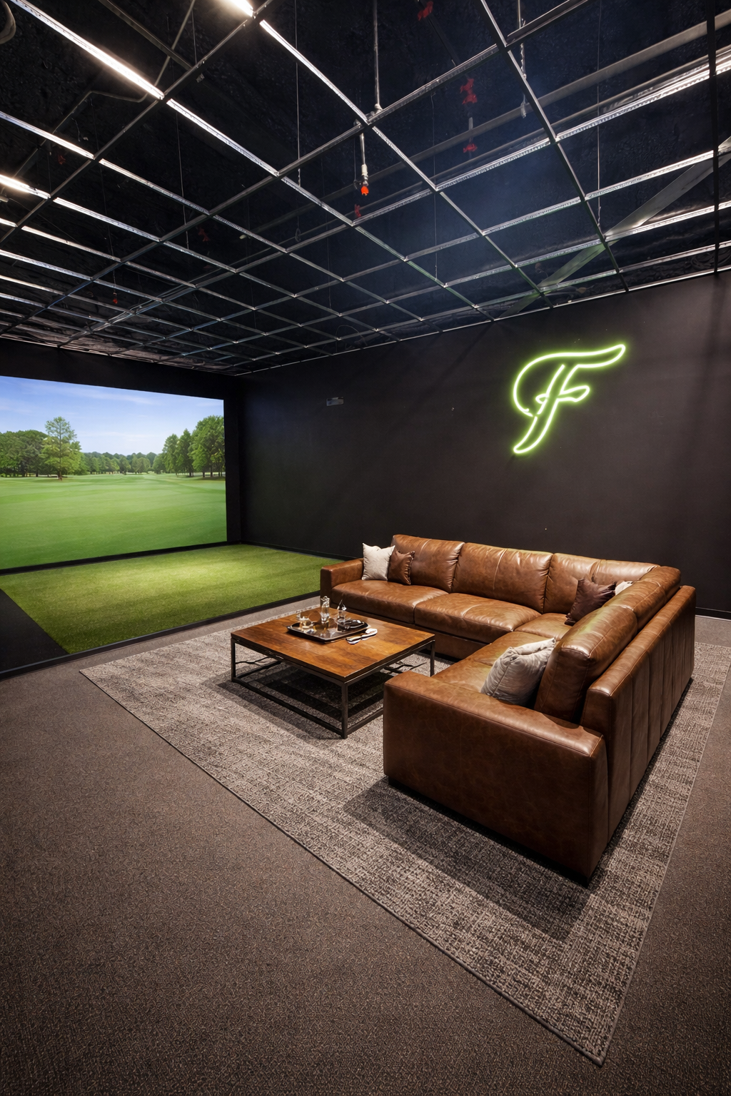 Indoor golf simulation room with a large TV screen showing a golf course, a brown leather sectional sofa with pillows, a wooden coffee table with drinks and snacks, a illuminated green neon letter sign on the black wall, and a ceiling with exposed black grid panels and track lighting.