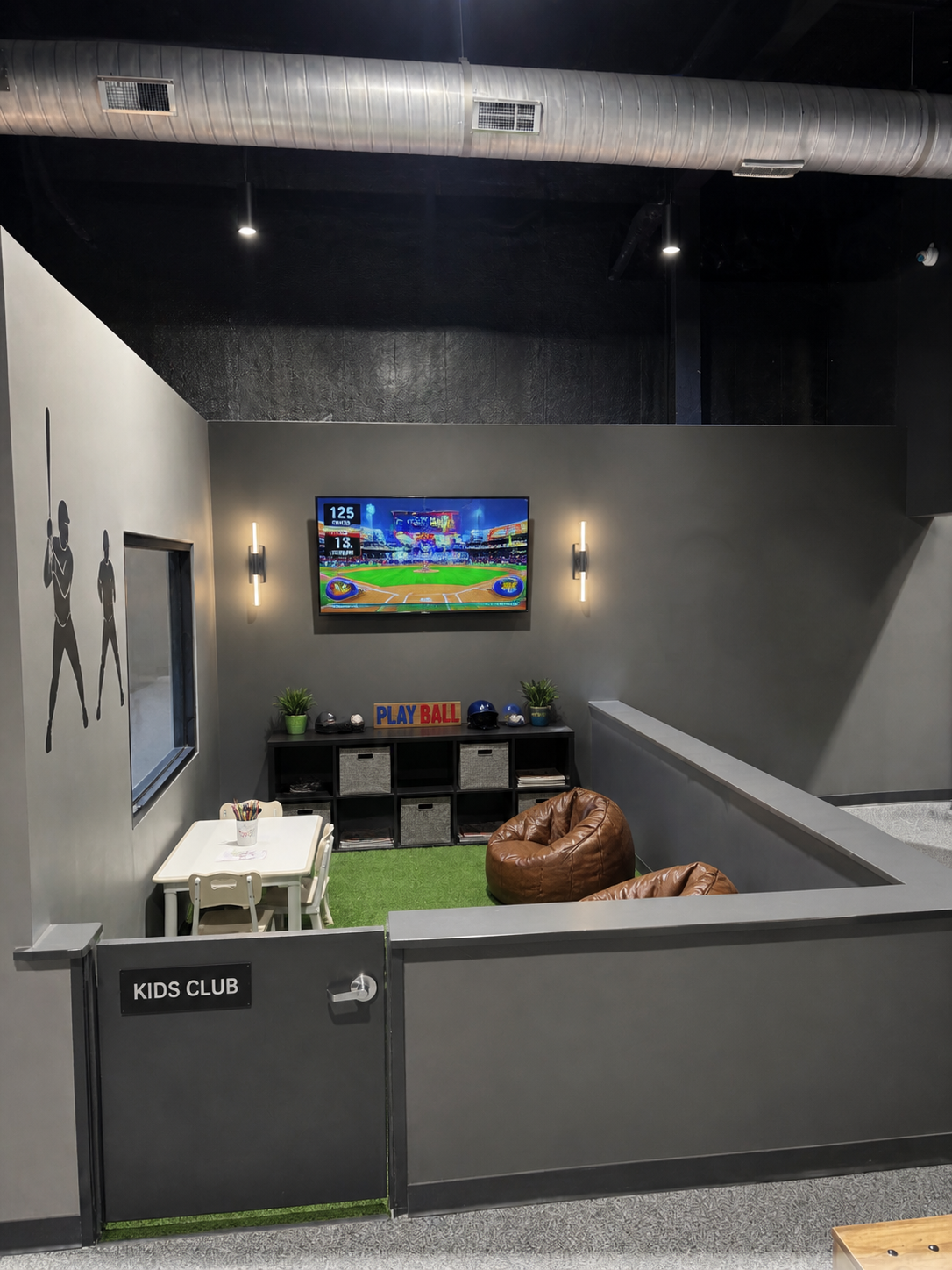 Children's indoor play area labeled 'Kids Club' featuring a small table with chairs, bean bag chairs, a wall-mounted TV showing a baseball game, shelves with toys and decor, and baseball-themed decorations.