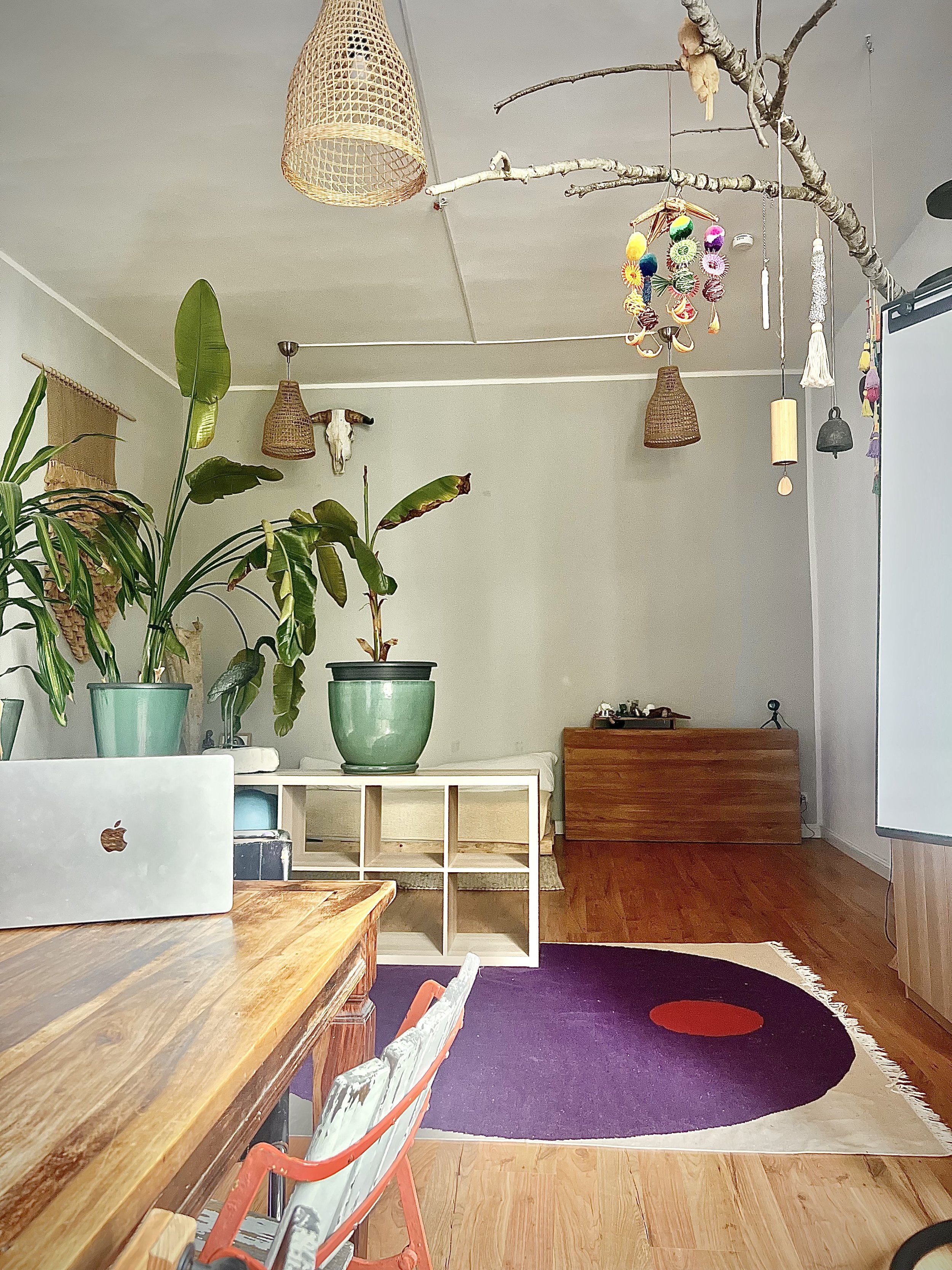 Gemütliches Wohnzimmer mit Holztisch, Pflanzen, buntem Teppich, hängenden Dekorationen und Tageslicht.