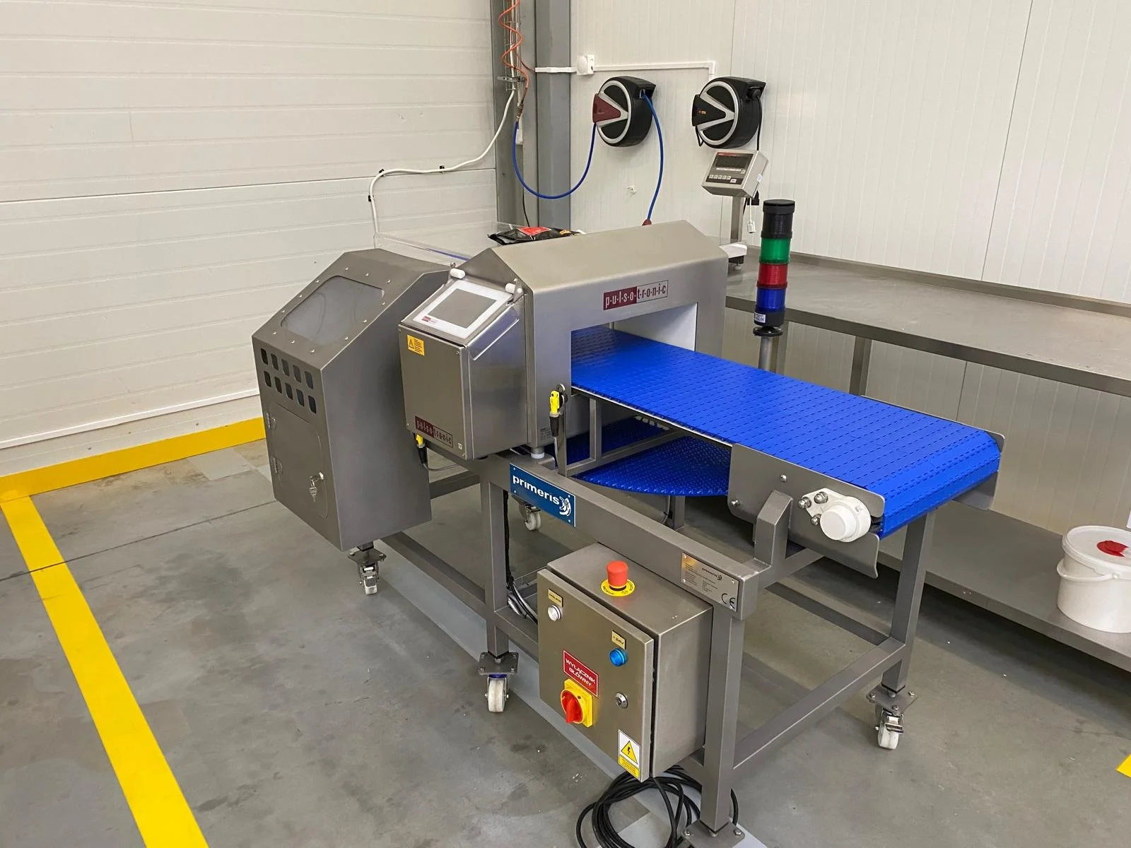 Industrial conveyor belt system with control panel and warning lights in a clean, gray-walled manufacturing or processing facility.