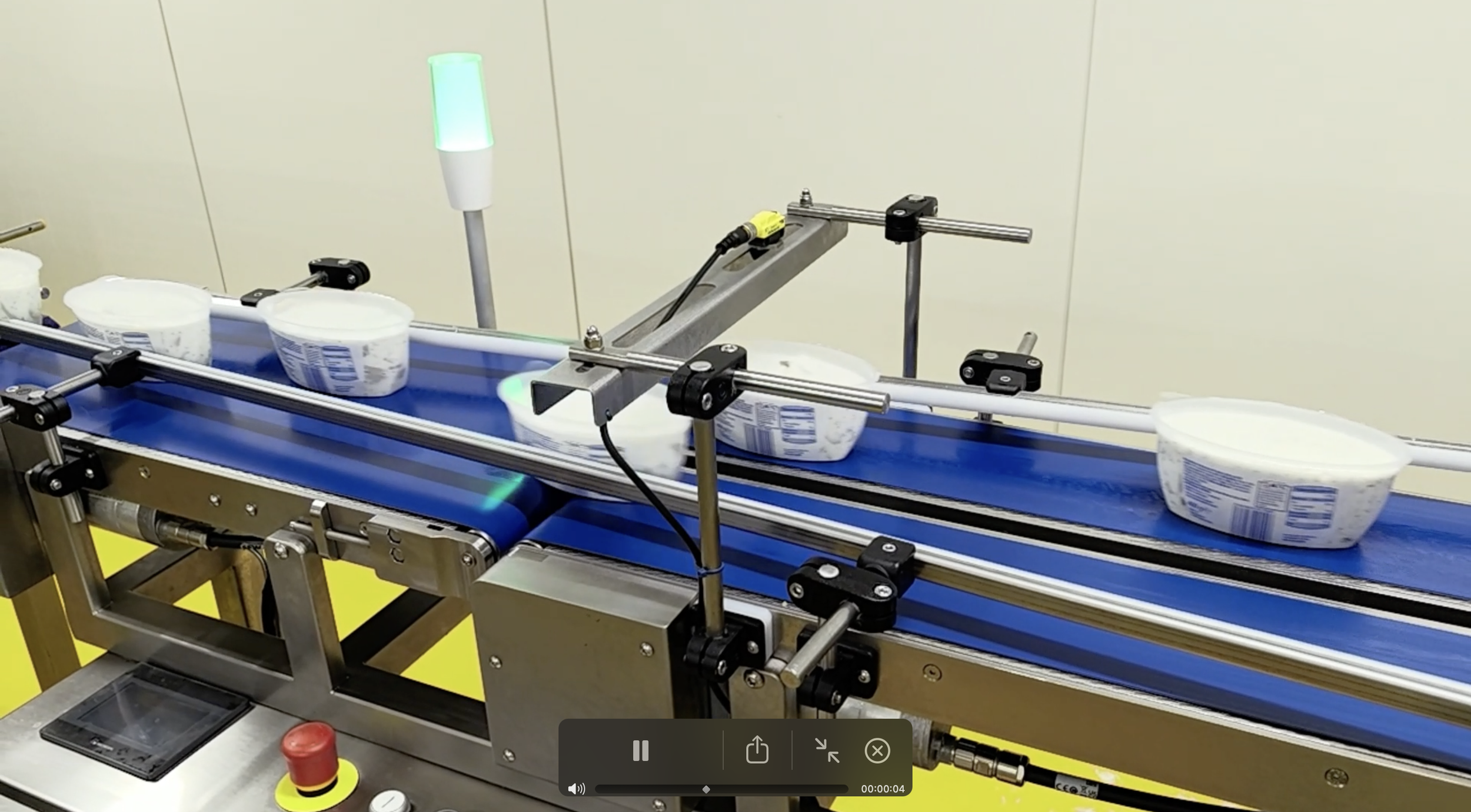Automated conveyor belt with three containers of yogurt moving along a factory production line, equipped with sensors and mechanical supports in a sterile manufacturing environment.