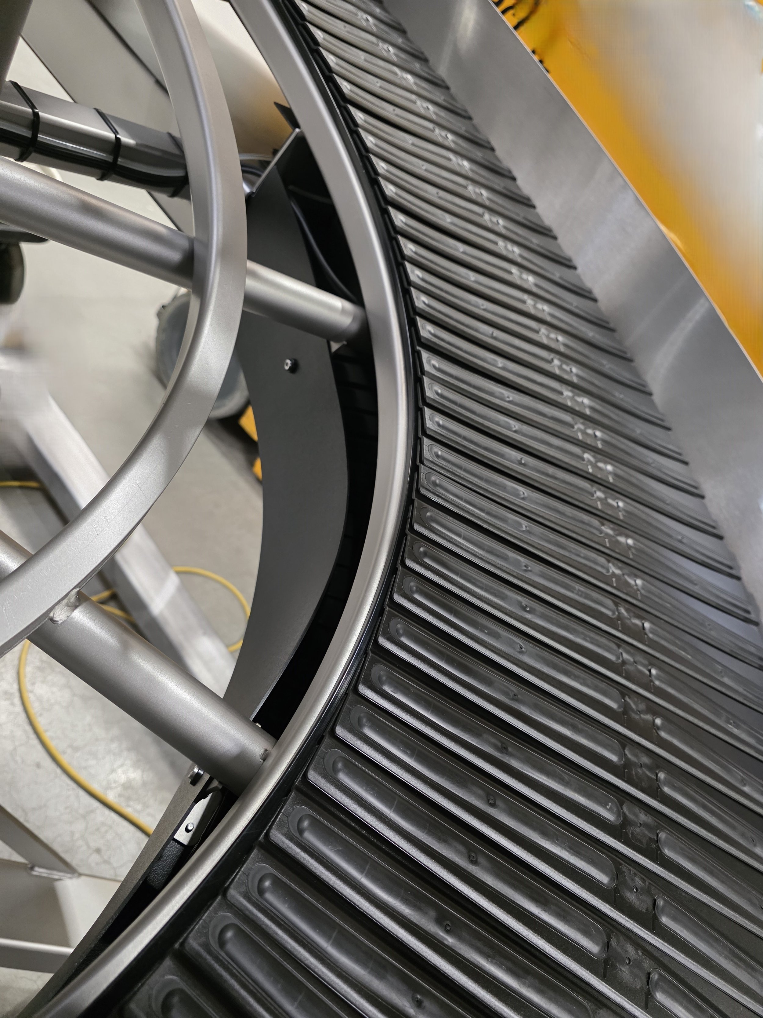 A close-up view of a large black conical device with a circular opening and a curved, segmented surface made of elongated rectangular sections. There are metallic support structures and a yellow cord in the background.