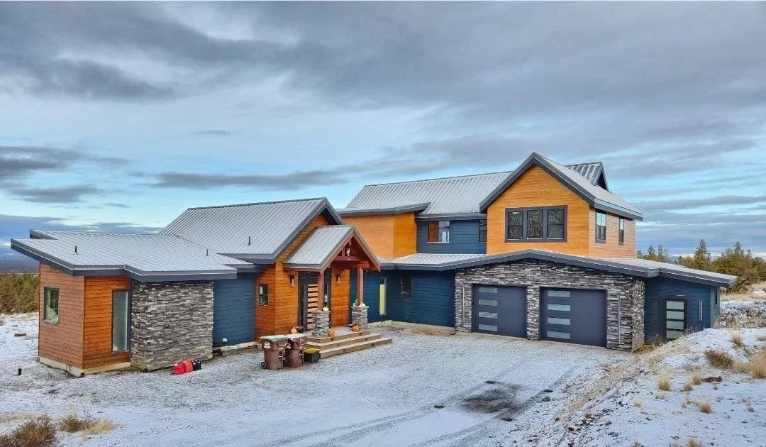 Modern two-story house with a mix of wooden, stone, and dark-colored exterior, multiple garage doors, situated in a semi-rural area with a mostly cloudy sky.