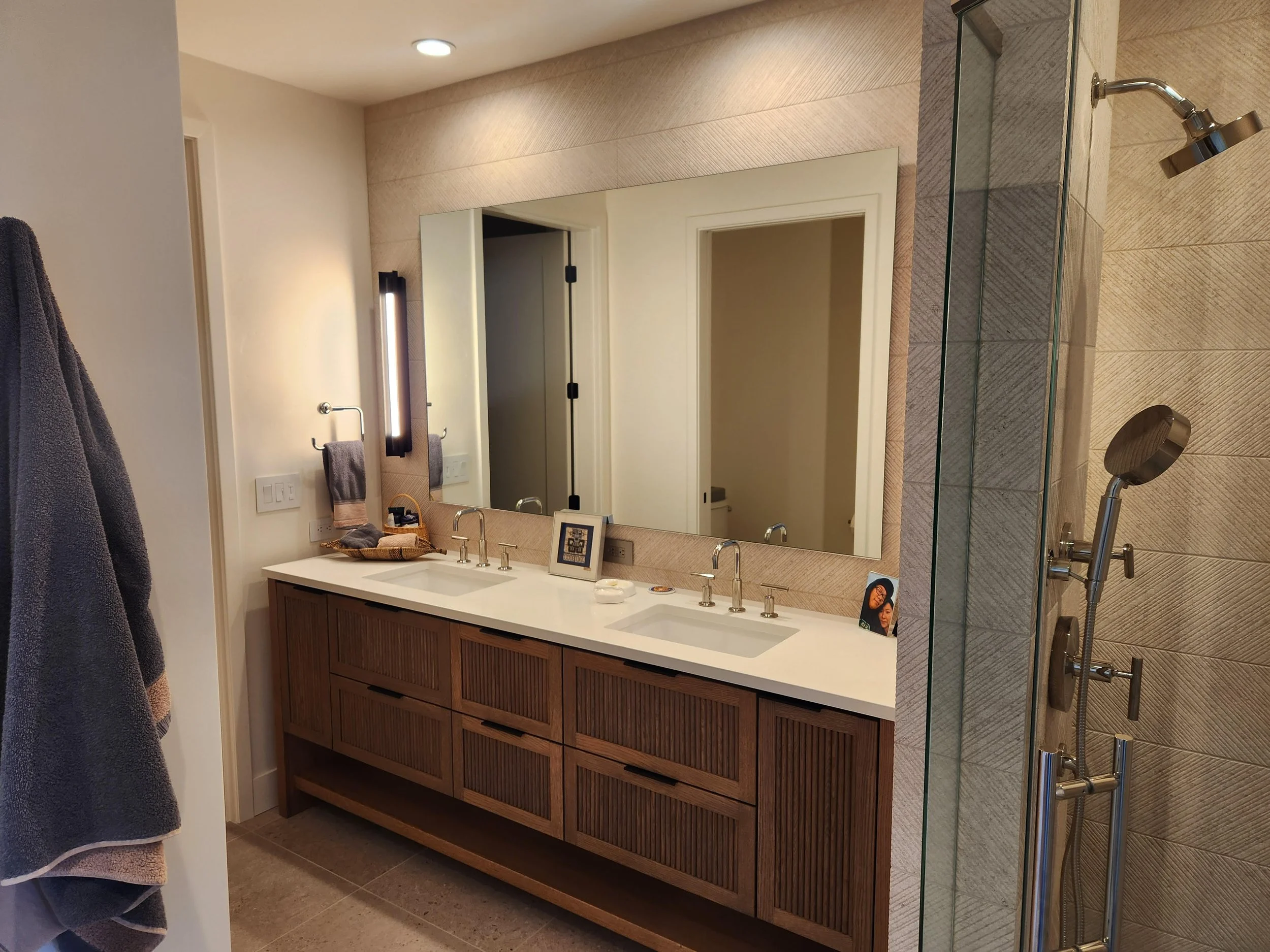 Bathroom with double vanity, large mirror, wall-mounted lights, towels, framed photo, and a glass-enclosed shower with multi-showerheads.