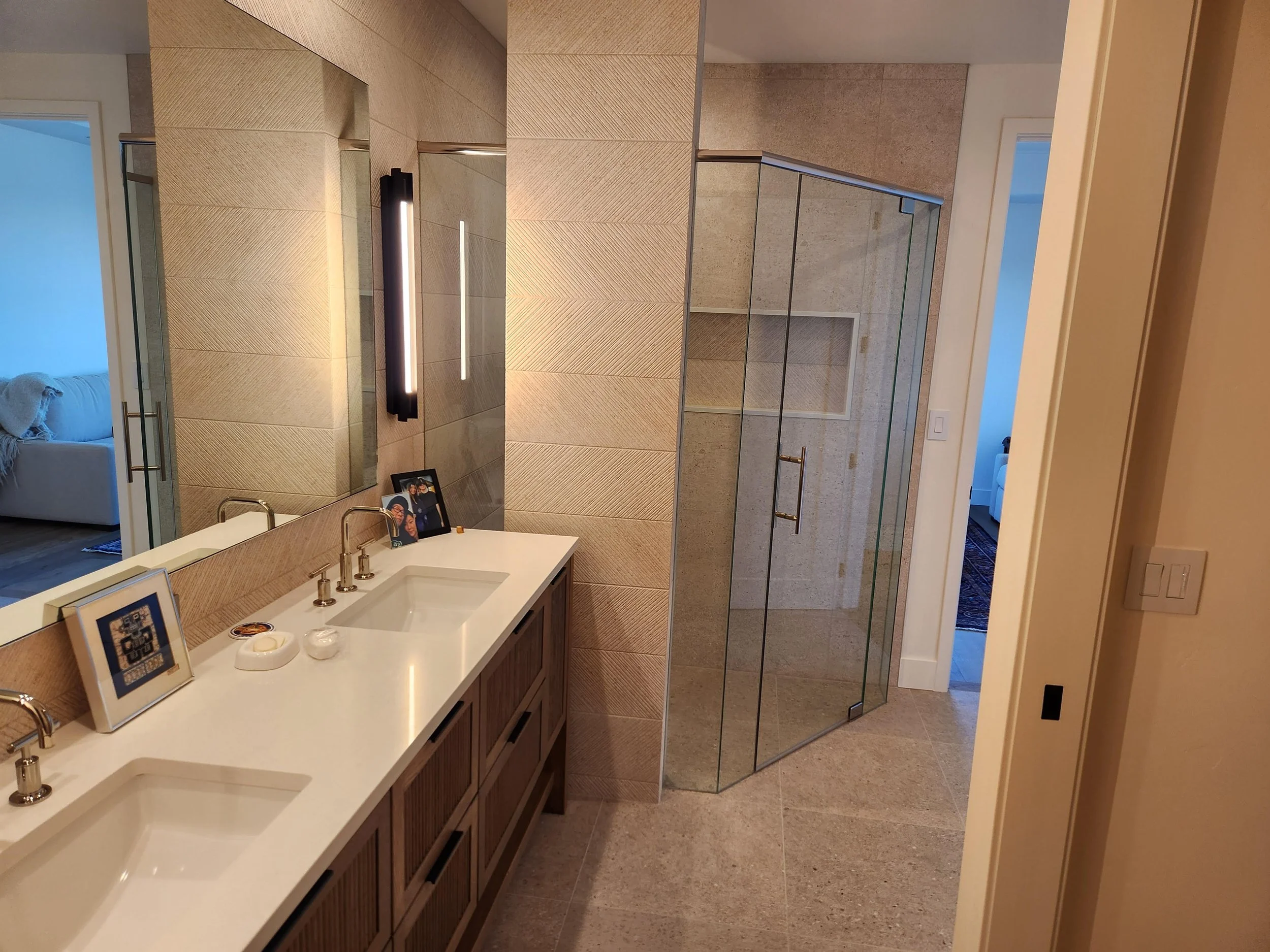 Bathroom with dual sinks, large mirror, framed photos, modern black wall-mounted light fixture, glass shower enclosure, beige tiles, and an open door leading to a living room.
