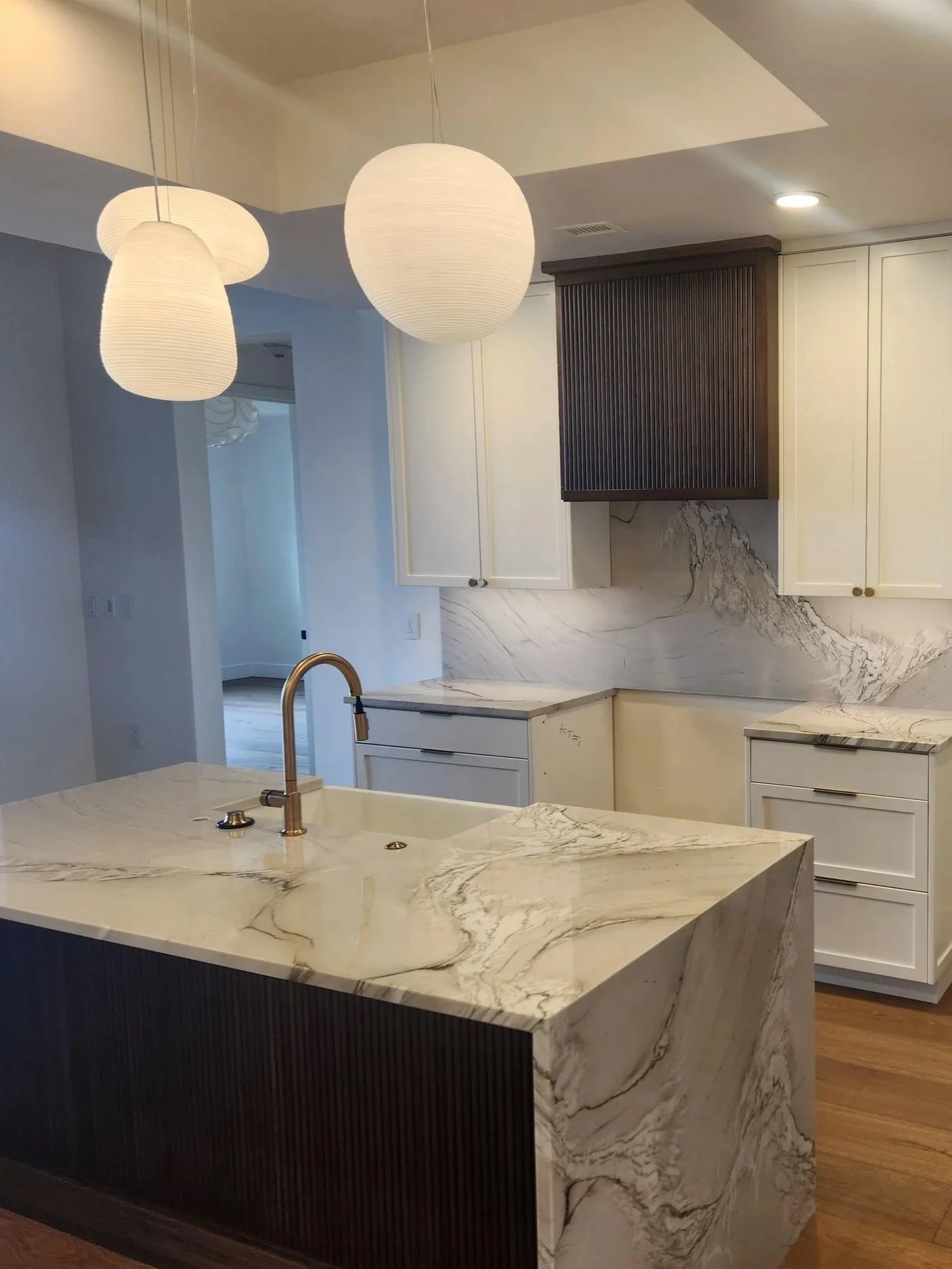 Modern kitchen with white marble countertops and island, white cabinets, black wood-paneled cabinet above stove, and pendant lights overhead.