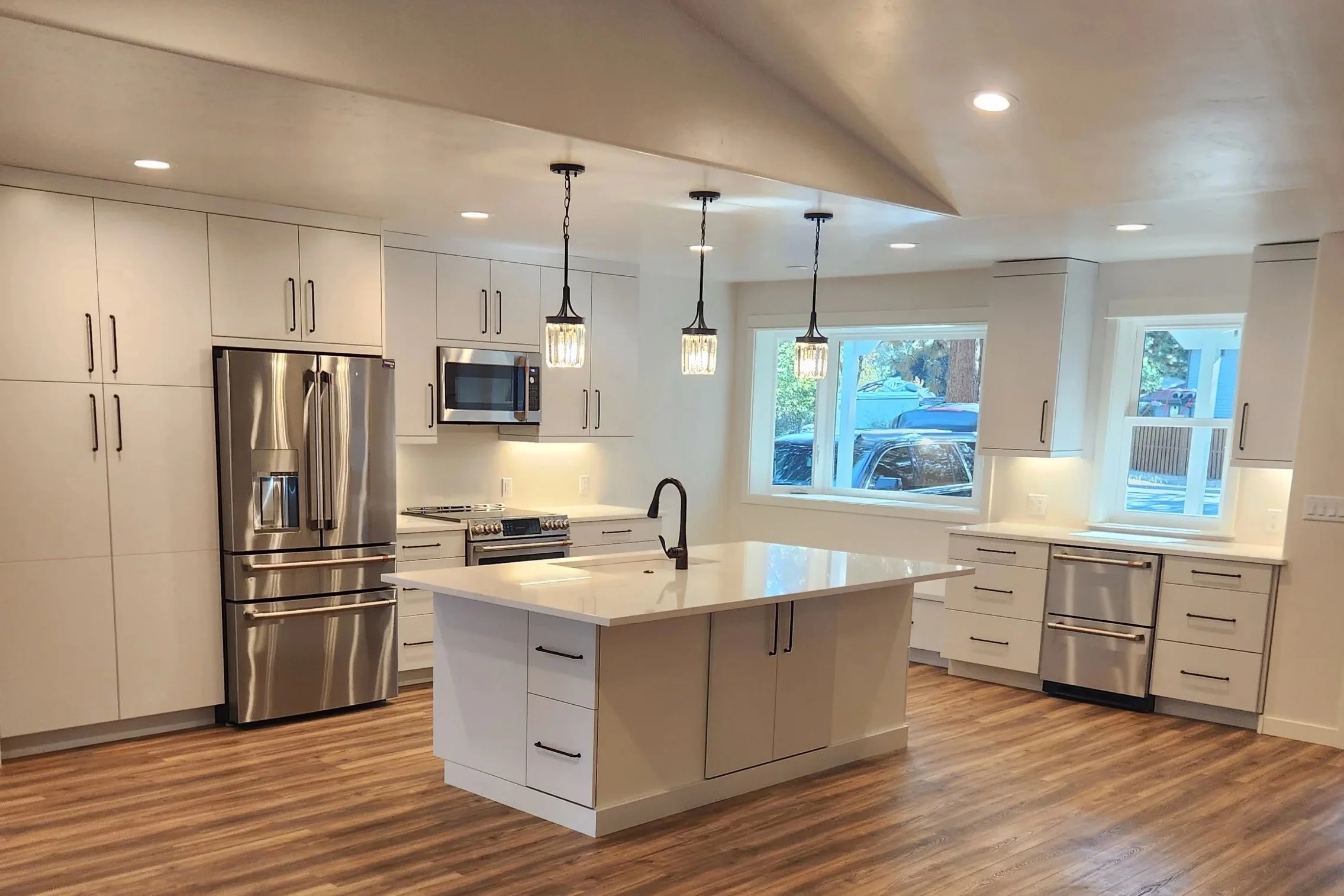 Modern kitchen with white cabinets, stainless steel refrigerator, microwave, and stove, center island with sink, hardwood floors, large windows, and pendant lighting.