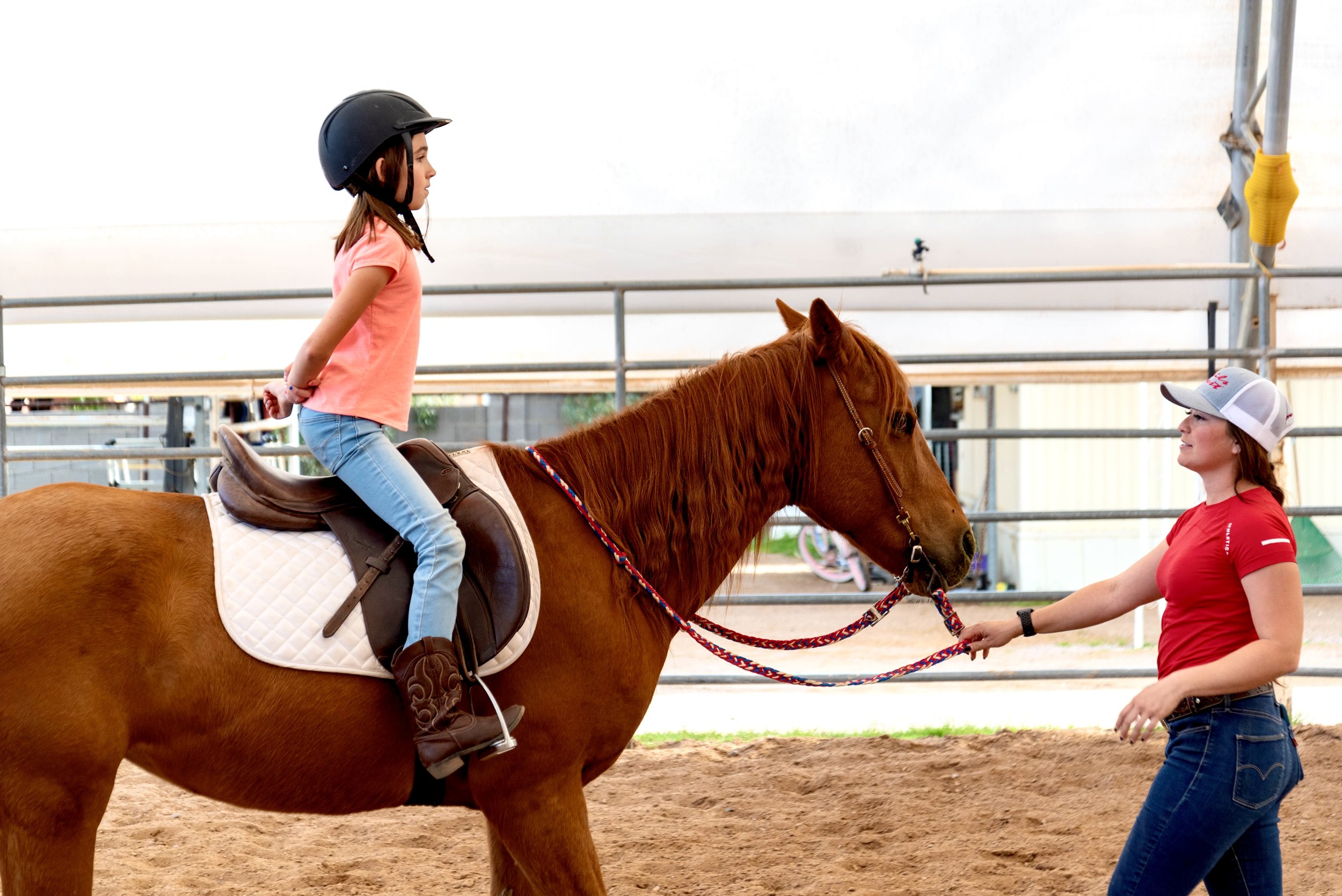 A girl wearing a black riding helmet and pink t-shirt riding a chestnut horse with a woman guiding the horse by holding its reins at a riding arena.