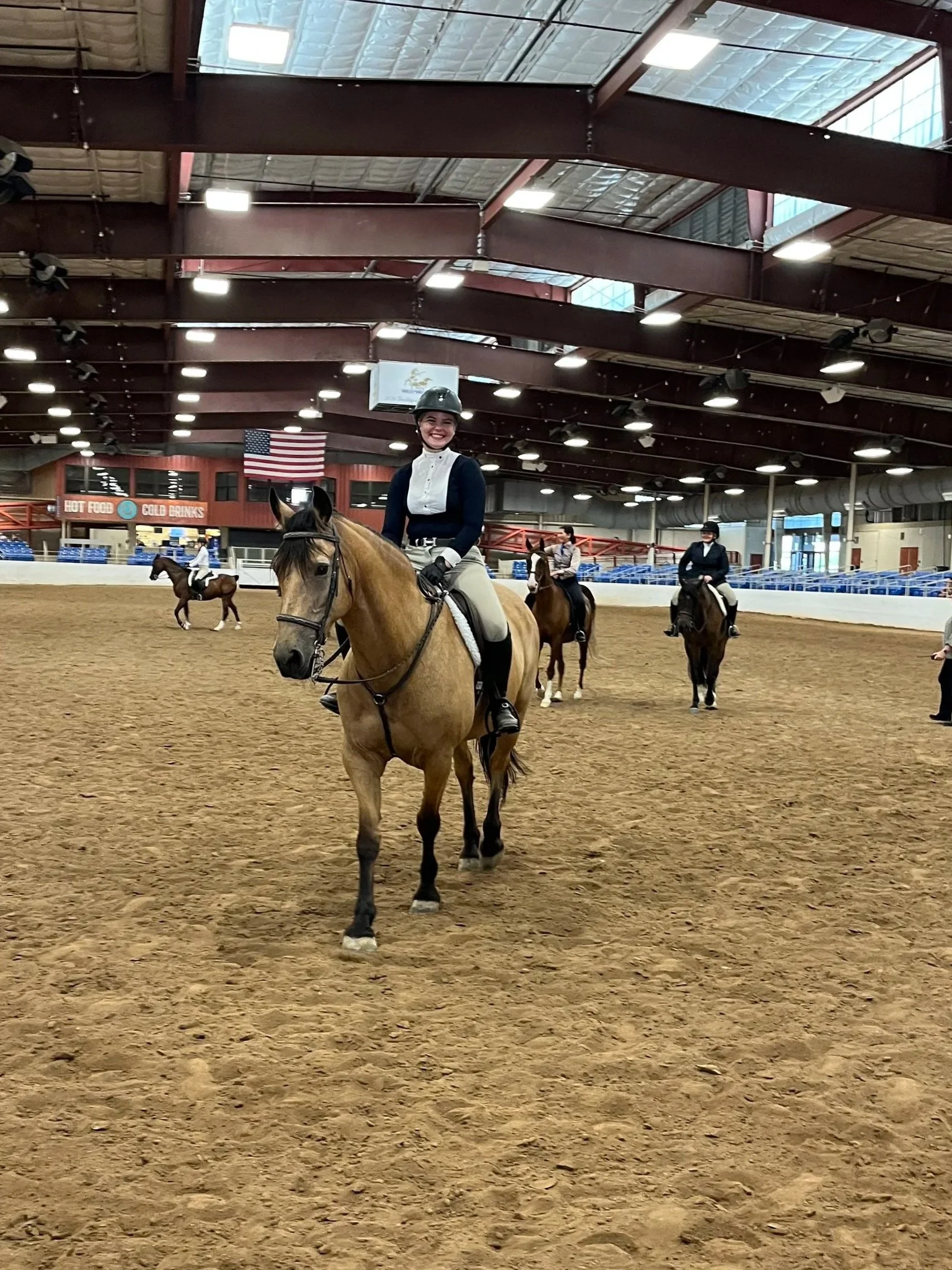 An indoor equestrian arena with riders on horseback. A smiling woman in a helmet and riding attire is in the foreground on a beige horse. In the background, other riders and horses are visible, along with American flags and signs for hot food and cold drinks.
