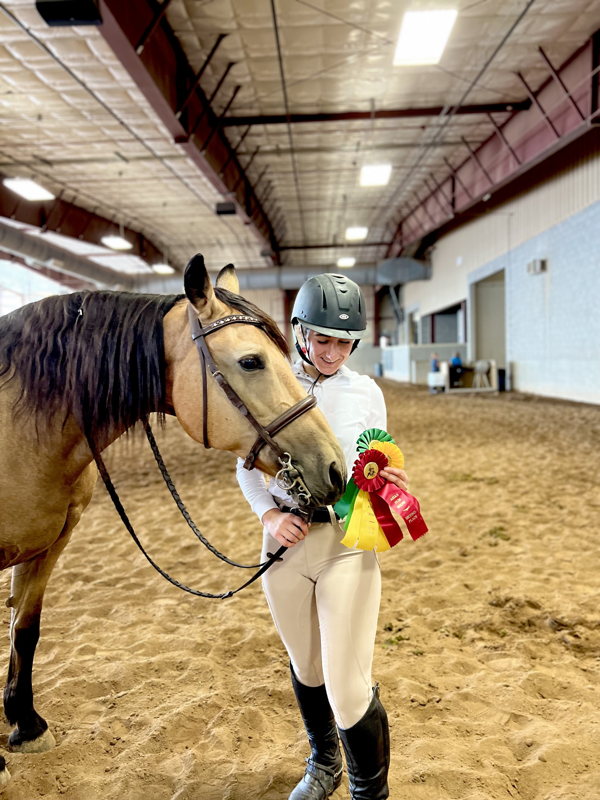 A woman in riding attire holding ribbons standing next to a horse in an indoor arena with a high ceiling.