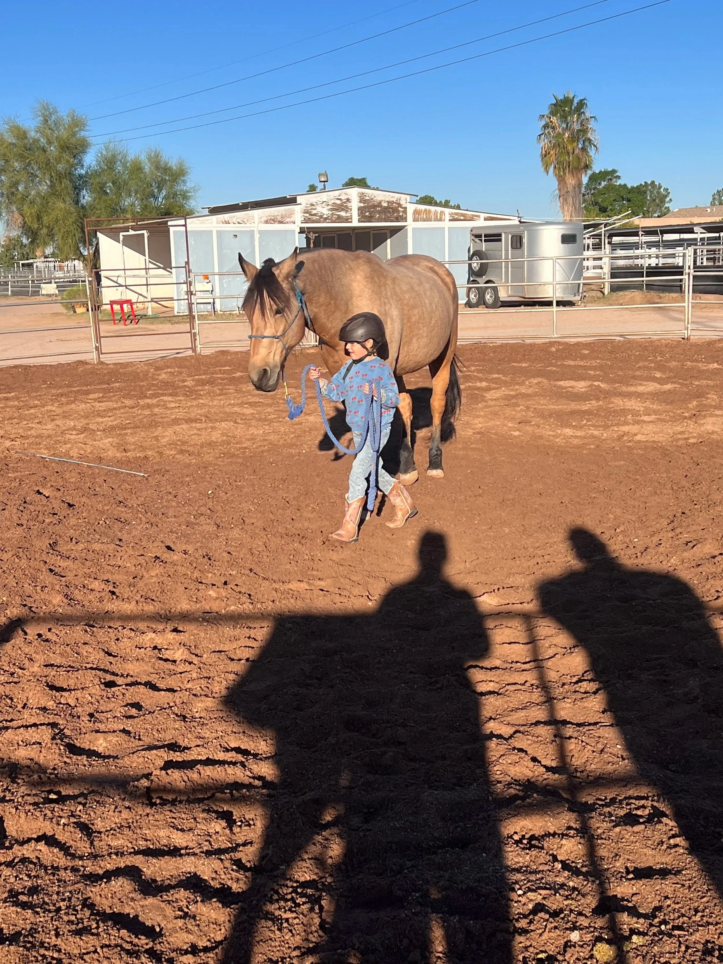 A young girl wearing a helmet and cowboy boots holding a lead rope connected to a brown horse with a black mane inside a dirt riding arena. There are trees, a building, and a trailer in the background, with clear blue skies.