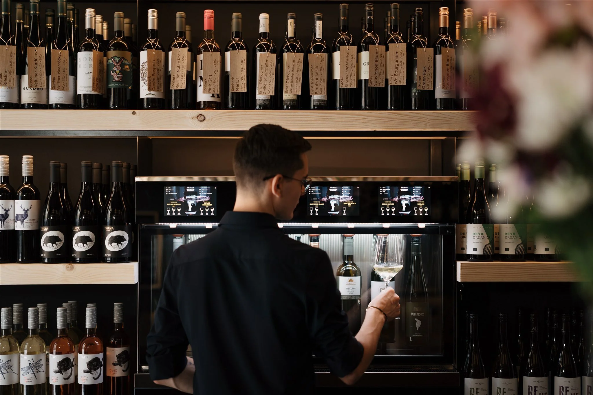 A person wearing glasses and a black shirt is pouring wine in a wine bar or shop. There are shelves filled with wine bottles behind them.
