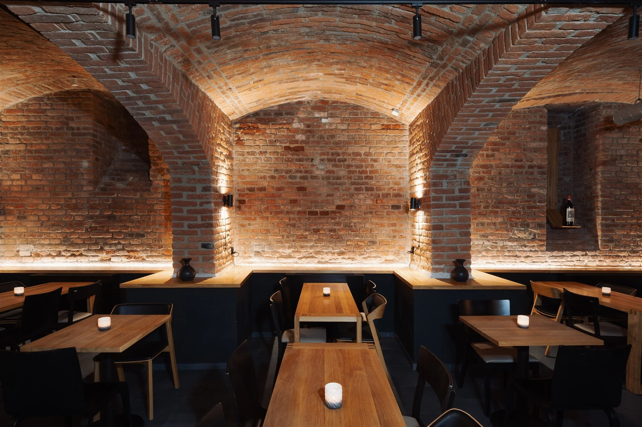 Interior of a restaurant with exposed brick walls and vaulted brick ceiling. Wooden tables with black chairs are arranged throughout, each table has a small lit candle. The overall ambiance is warm and rustic.
