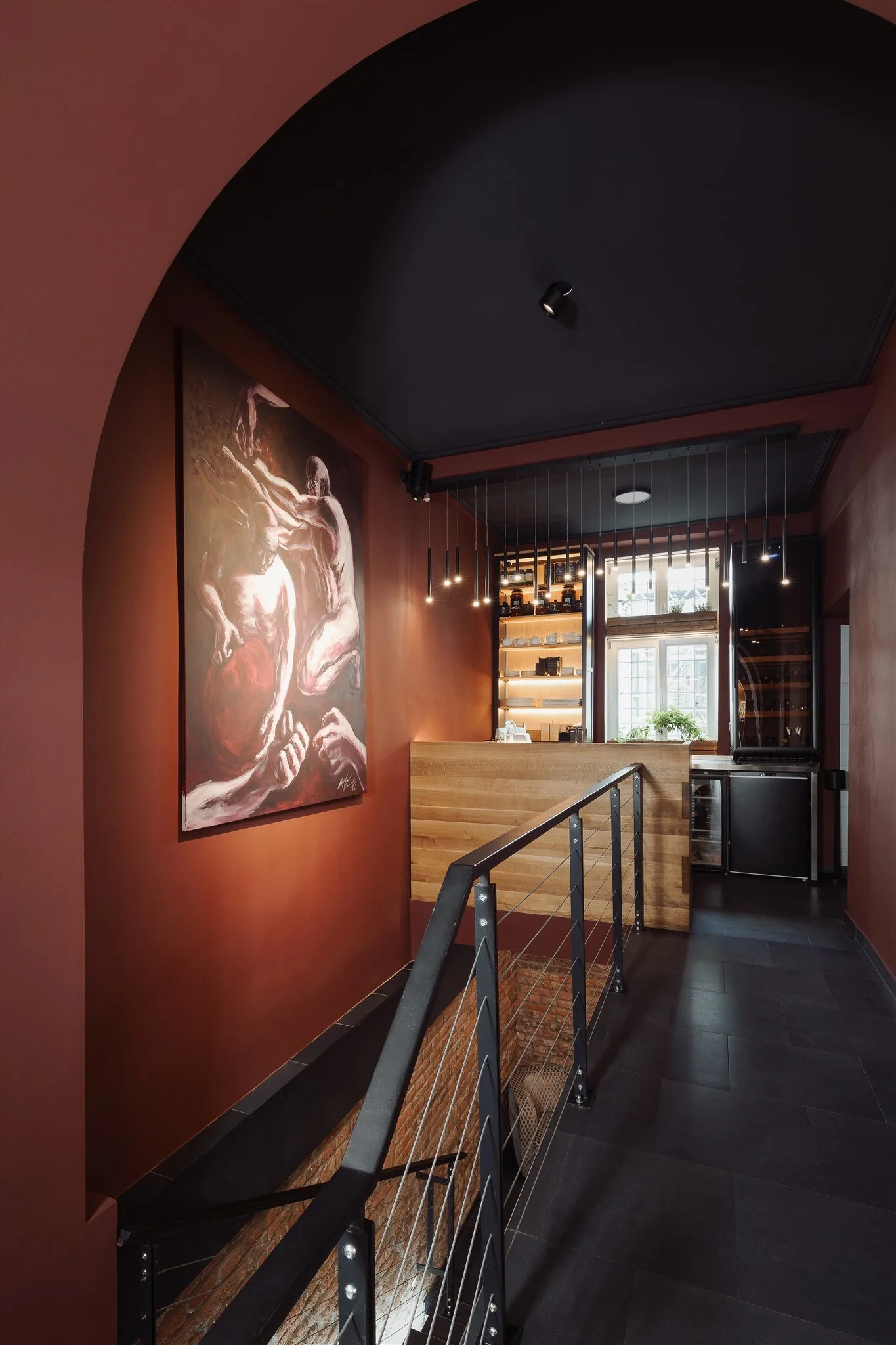 Interior of a modern cafe or restaurant with a dark ceiling, large abstract art on a reddish wall, black flooring, and a metal railing overlooking a lower level with shelves and large windows letting in natural light.