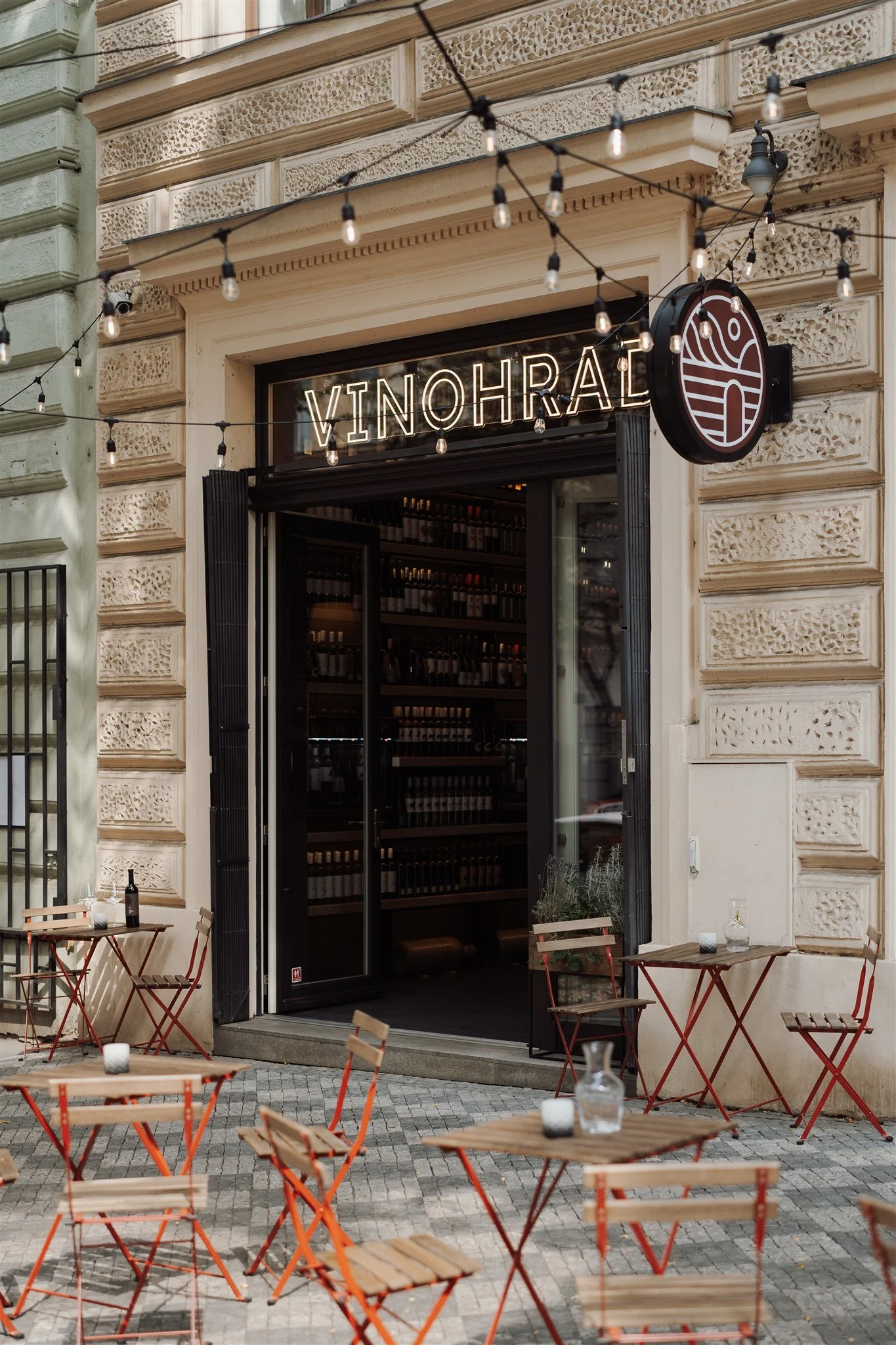 Outdoor cafe with empty wooden tables and chairs, string lights overhead, in front of a wine shop named VinoHRÁL with an open door revealing wine bottles inside.