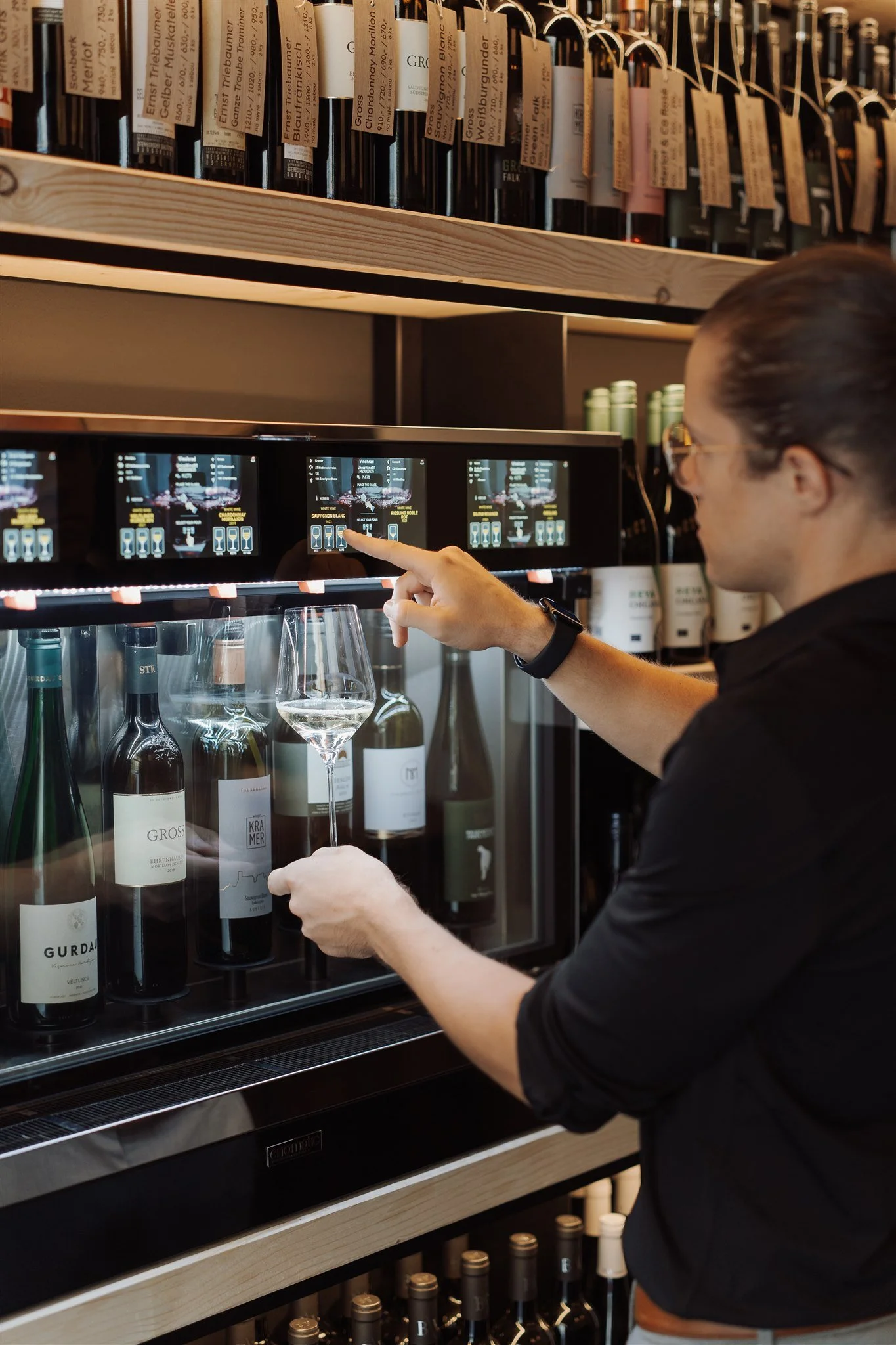 A person with glasses and dark hair is selecting a bottle of wine from a wine cooler. There are wine bottles on shelves and labels visible in the cooler and above.