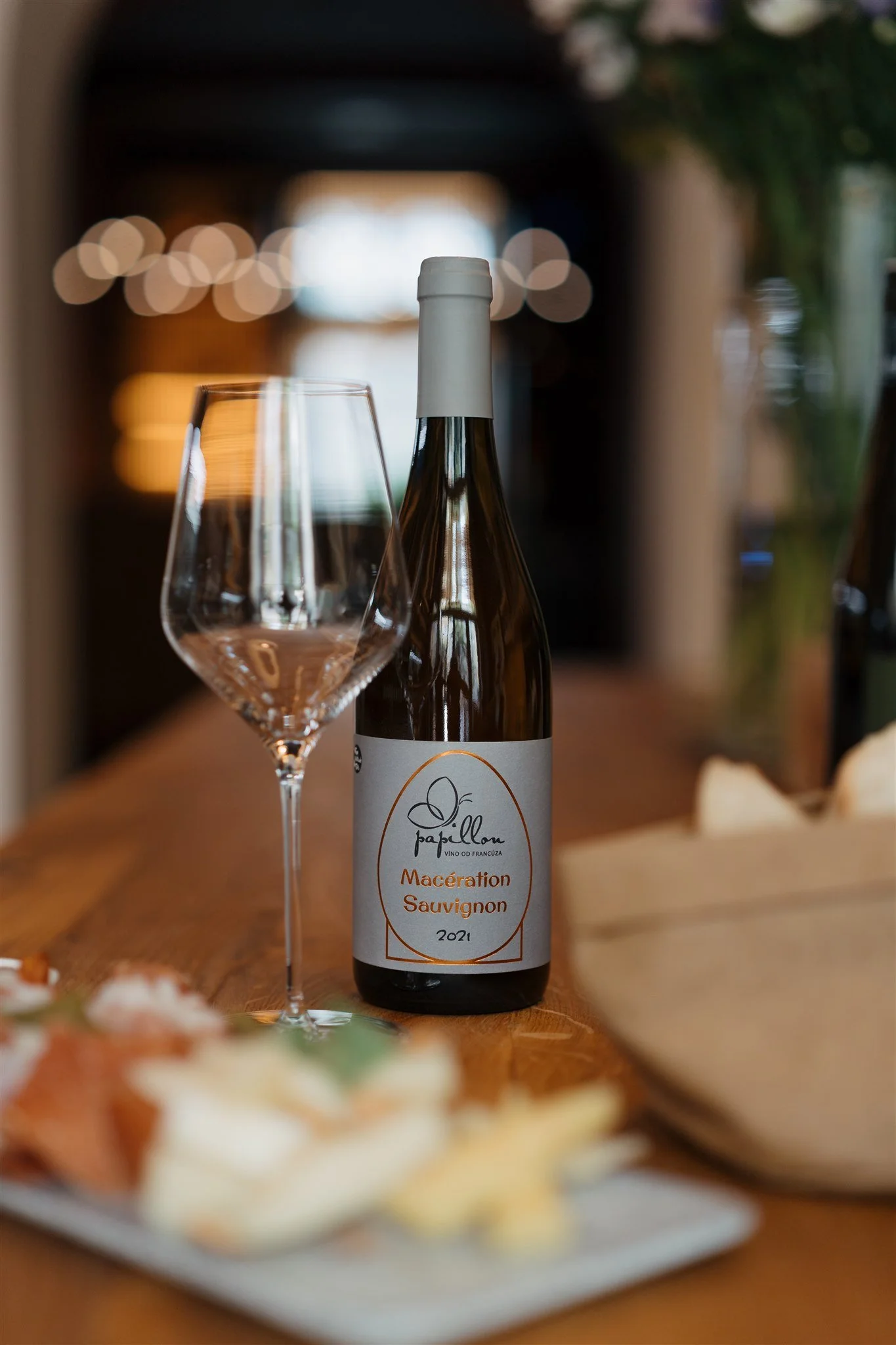 A bottle of wine labeled Maceration Sauvignon with a glass beside it on a wooden table, with blurred warm lighting and decor in the background.
