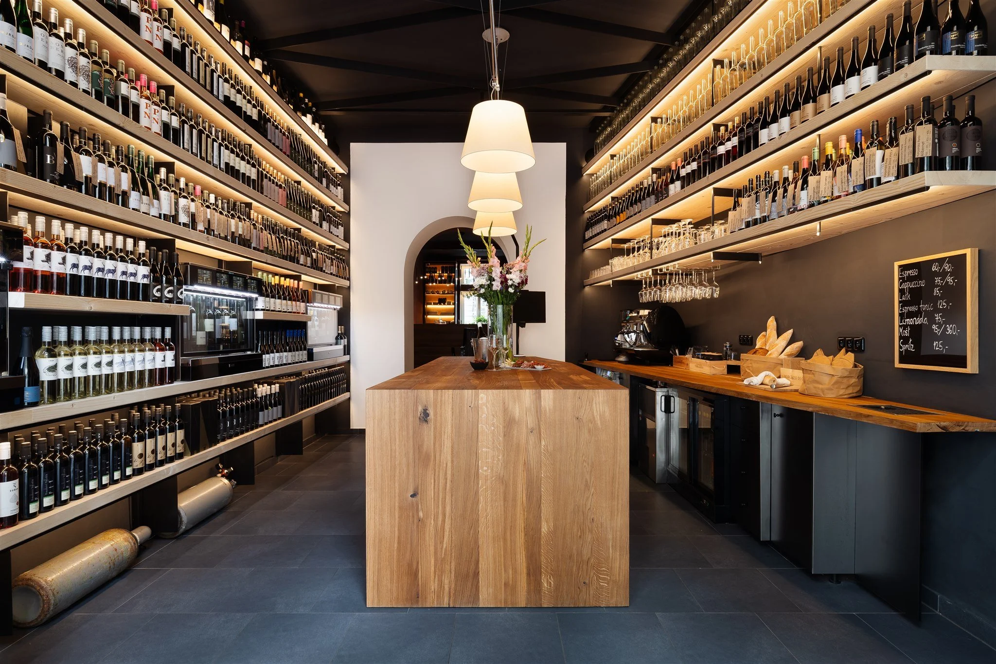 A wine shop interior with wooden shelves filled with wine bottles, wine glasses hanging, a central wooden counter with flowers, and a black chalkboard menu on the right side.