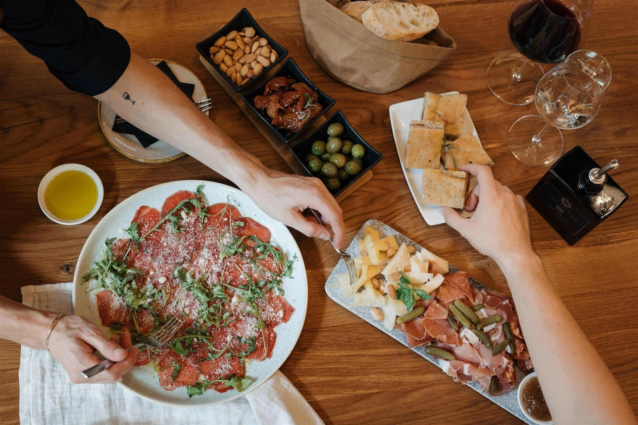 People sharing a meal with various appetizers and drinks on a wooden table, including a pepperoni pizza, bread, olives, roasted meat, charcuterie, and glasses of red and white wine.