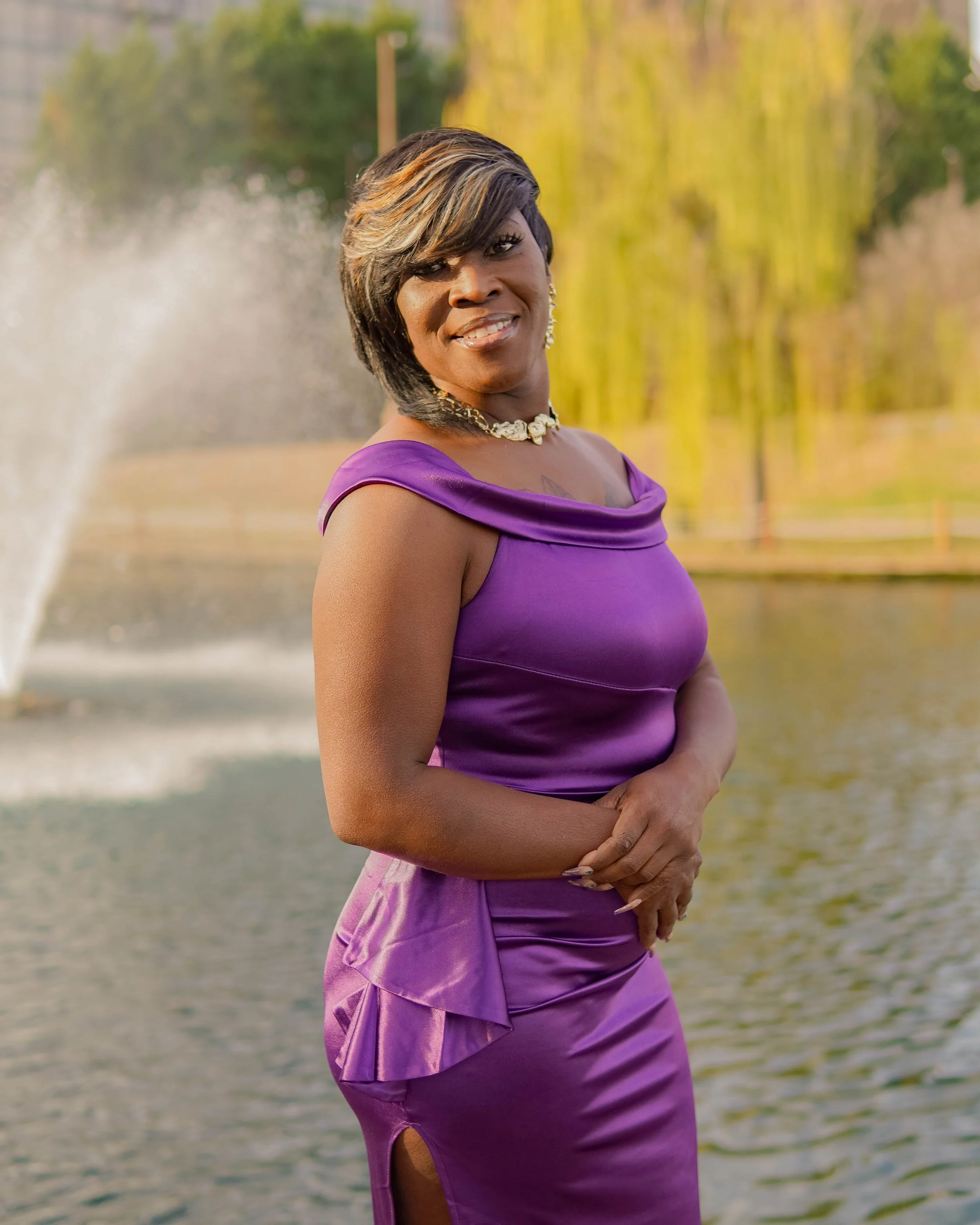 A woman in a purple dress standing near a pond with a fountain and trees in the background.