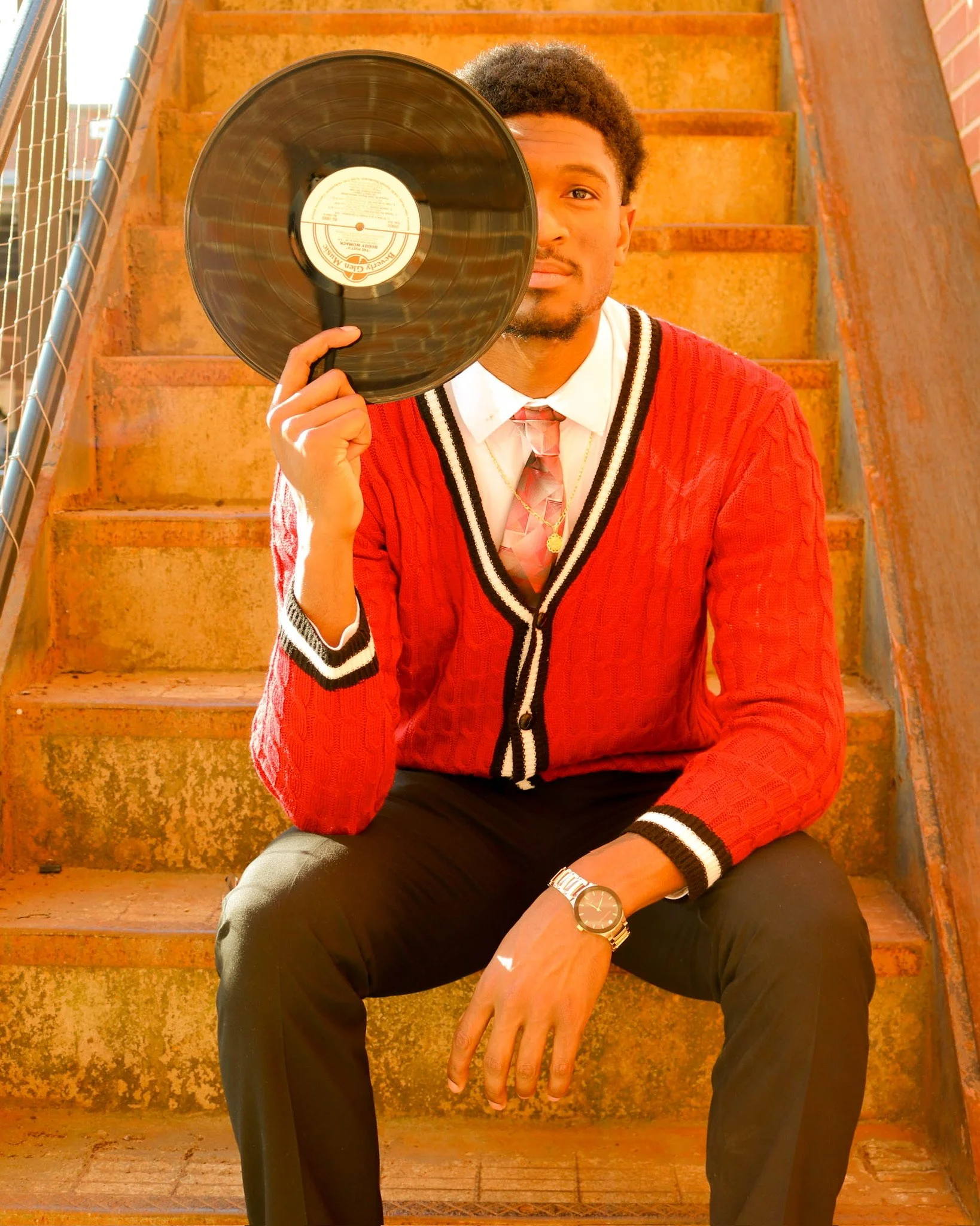 A young man sitting on outdoor stairs, holding a vinyl record in front of his face, with one eye peeking over it, wearing a red cardigan, white shirt, patterned tie, black pants, a wristwatch, and gold jewelry.