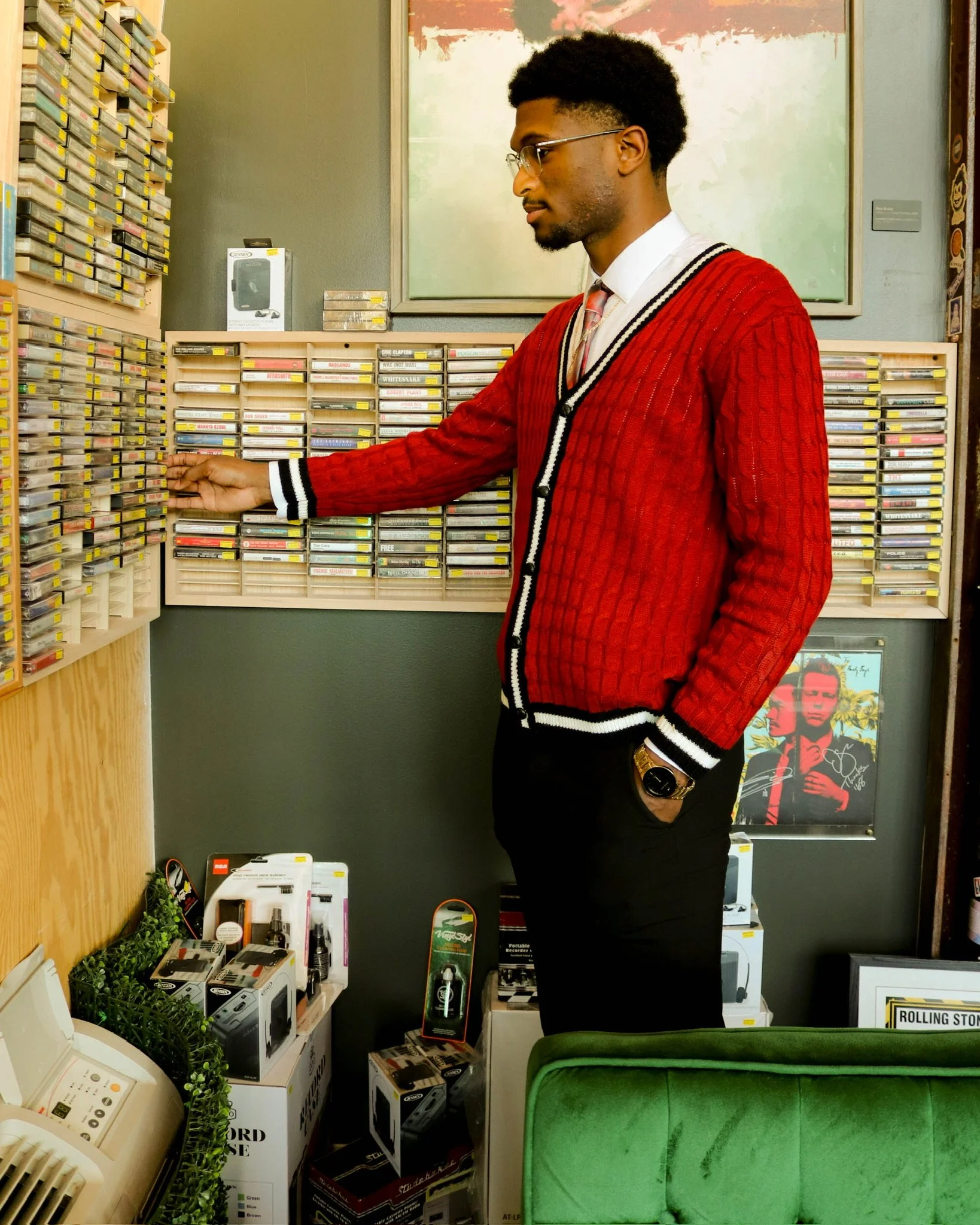 A young man in glasses, a white shirt, and a red sweater with black and white details, standing in front of a wall of cassette tapes, selecting one. The setting appears to be a record or gadget store with various electronic items on display.